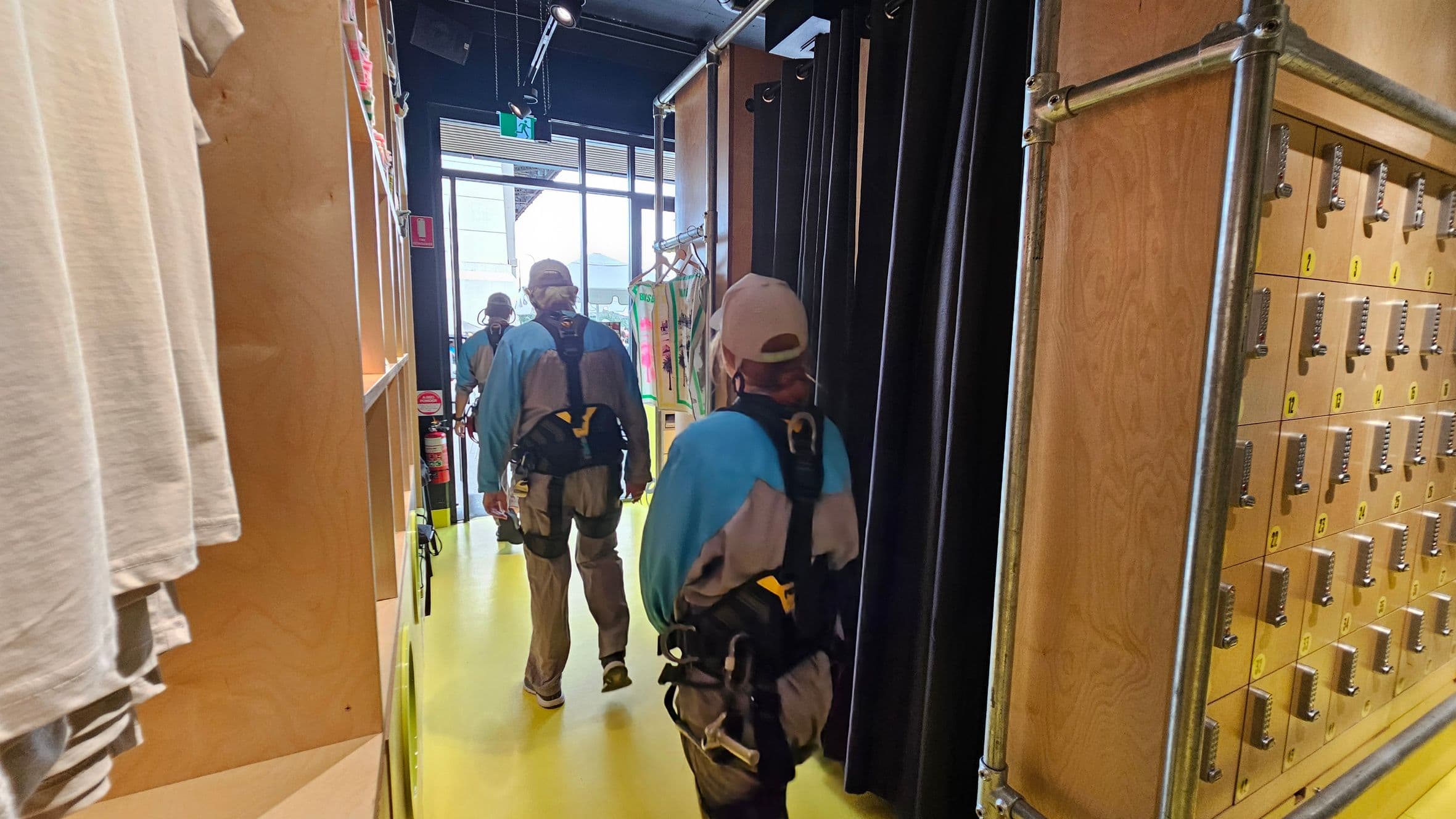 A narrow corridor with a bright yellow floor. On the left, there are shelves with folded clothes and a hanging white T-shirt. On the right, a wall of small lockers with combination locks. In the centre, several people wearing harnesses and helmets walk towards a glass door at the end of the corridor. The ceiling is dark with visible pipes and lighting fixtures.