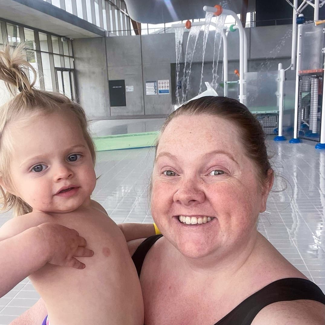 A smiling woman holds a baby in a bright indoor pool area.