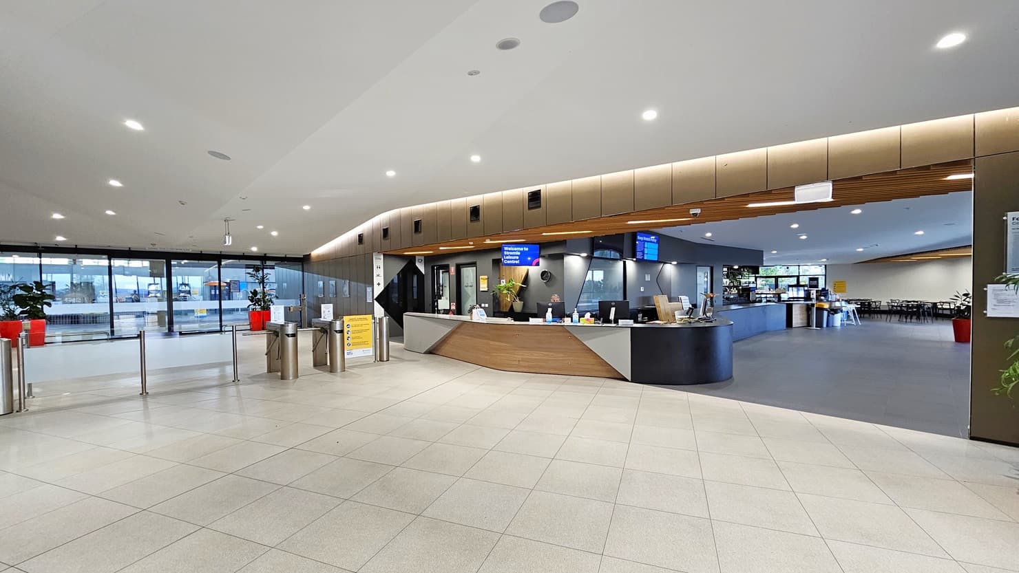 An automatic glass door, adorned with posters, provides access to the facility. Red planter boxes with indoor plants are positioned on both sides. Cushioned benches are on the left. The left wall features a brown wall with a flat-screen TV and retractable banners. To the right is the reception desk, featuring a brown and white front. A flat-screen TV is mounted from the ceiling. In the middle is a barrier gate. The ceiling features a white with scattered LED lights.