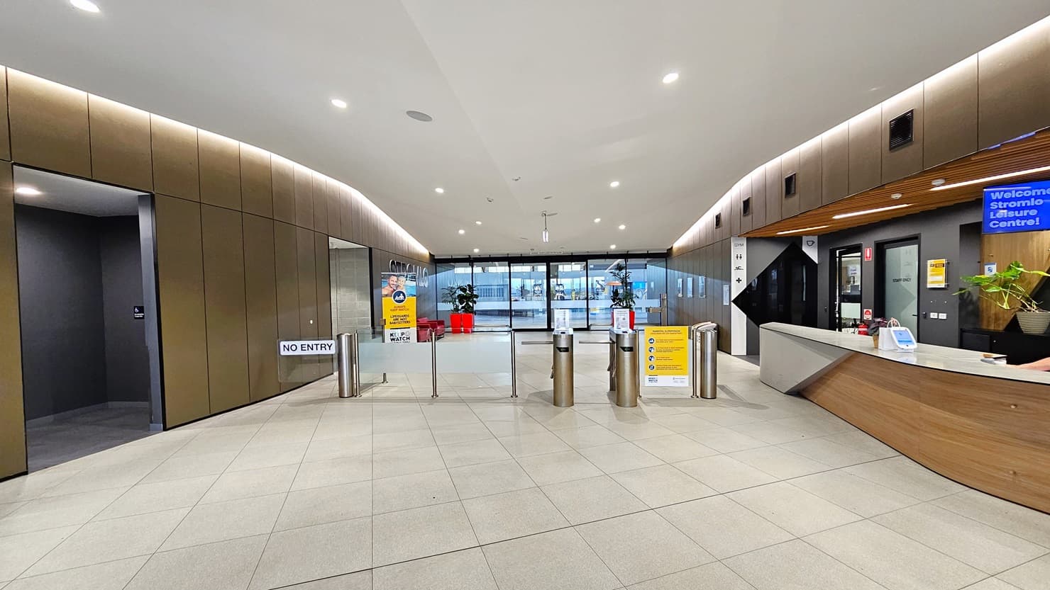 To the right is the reception desk, sporting a brown, white, and black front. A ceiling-mounted flat-screen TV is visible. In the middle is a turnstile gate. Further back, the main entrance is adorned with two red planter boxes and free-standing banners. On the left is an accessible bathroom. The reception desk extends into the corridor, leading to a seating area with chairs and tables on the right. The ceiling features a combination of white and brown with scattered LED lights.