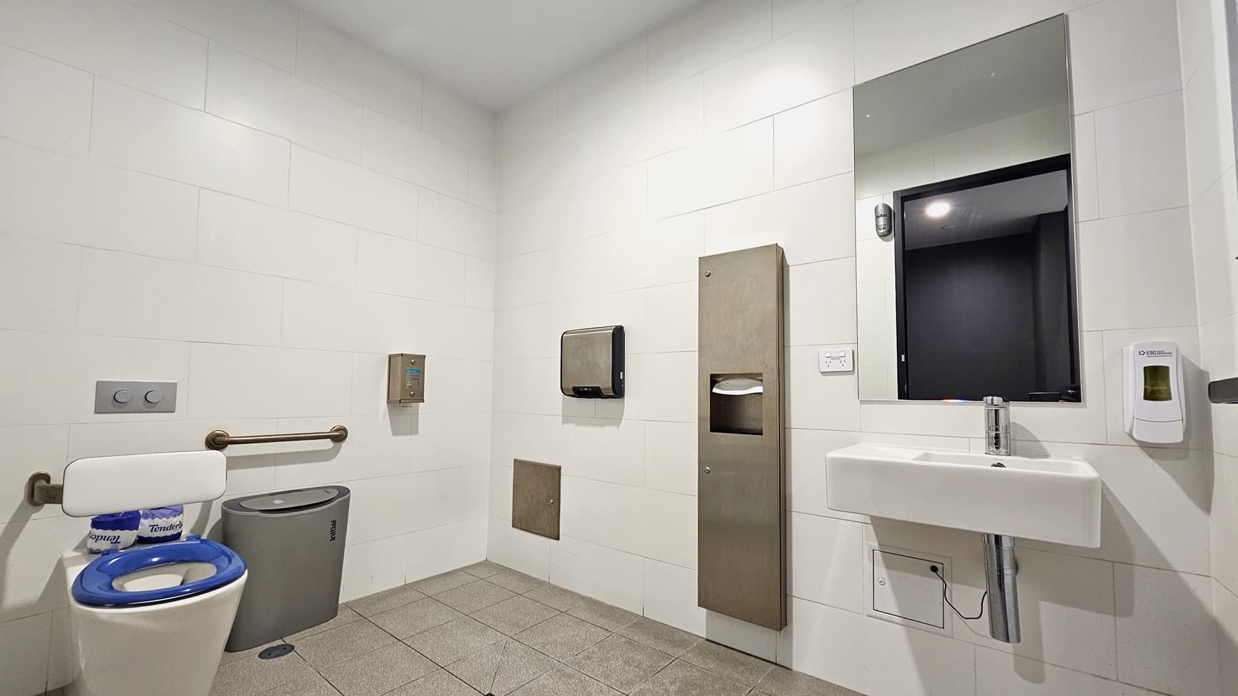 In the accessible bathroom, a toilet bowl with handrails is situated to the right, accompanied by a nearby rubbish bin. Adjacent to it, on the right, there is a wash basin, a mirror, and a soap dispenser. Additional features include a tissue dispenser and a hand dryer. The floor is covered in grey tiles, while the ceiling and walls are uniformly white.