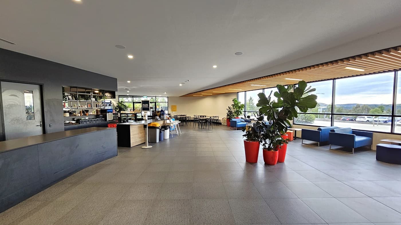 To the left is the extended reception desk and the café, featuring a sanitizer in the centre. The face exhibits a brown timber design. In the middle, there are pots of indoor plants. On the right, couches are positioned near the glass wall, and further in, there are movable plastic chairs and tables. Wooden planks adorn the ceiling on the right. The floor is white with LED lights, and grey tiles cover the floor.