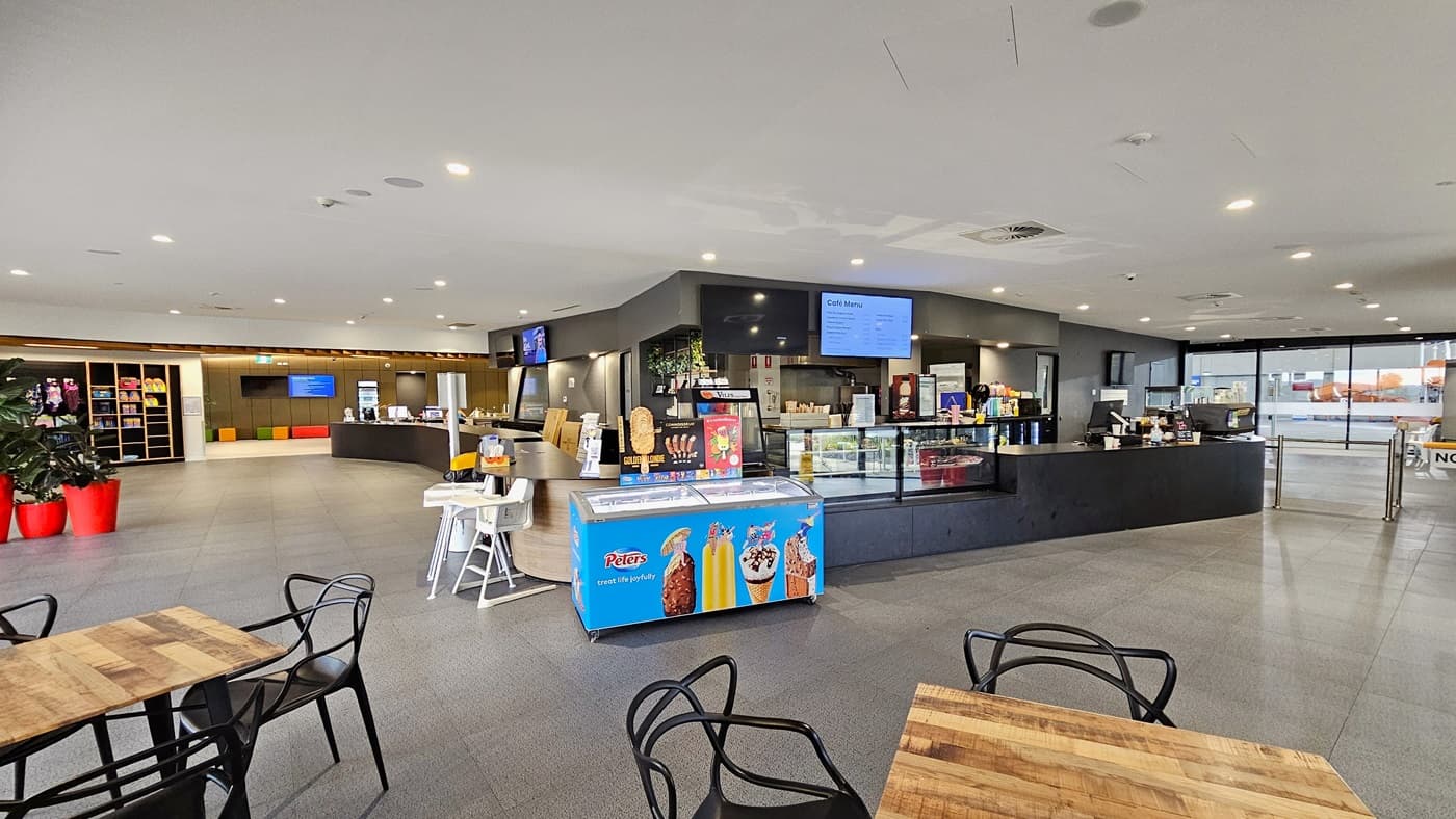 In the centre is the café with a black counter desk and a glass food display rack. A flat-screen TV is mounted on the wall, and an ice cream freezer is placed in the corner. On the left, a table holding flyers is situated, and further left, indoor plants in bright red pots are displayed along with a storage rack showcasing retail items. Movable plastic chairs and tables are arranged in front of the café. To the right is a turnstile gate.