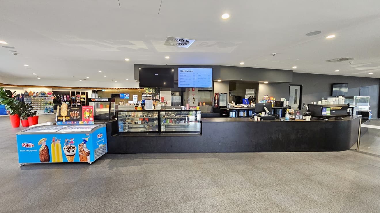 In the centre is the café with a black counter desk and a glass food display rack. A flat-screen TV is mounted on the wall, and an ice cream freezer is placed in the corner. On the left, a table holding flyers is situated, and further left, indoor plants in bright red pots are displayed along with a storage rack showcasing retail items. To the right is a turnstile gate. The floor is white with LED lights, and grey tiles cover the floor.