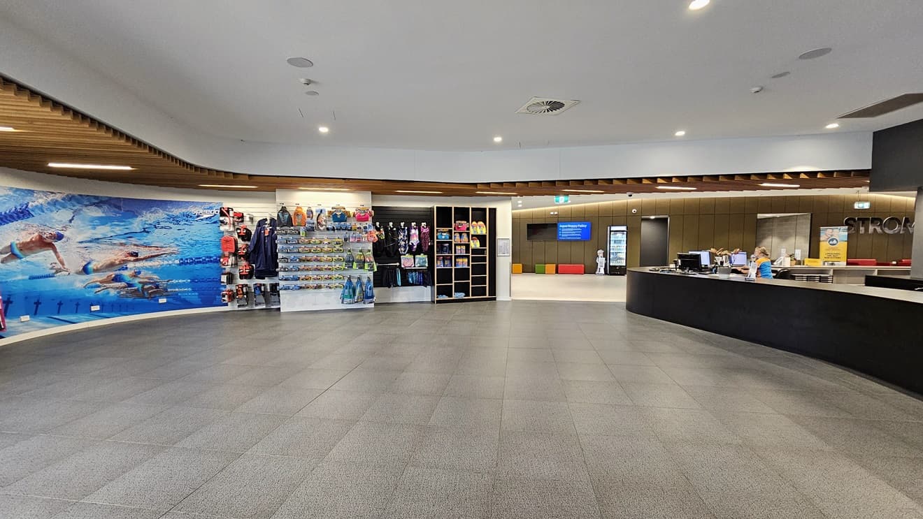 In the middle is a retail area displaying goggles, swimwear, and swimming accessories. To the right is the reception desk featuring an extensive black desk. To the left, the wall is adorned with a life-size poster of swimmers underwater. The ceiling has a cove design with wooden plank accents and LED lights. Grey tiles cover the floor. Further left, the brown wall holds flat-screen TVs, banners, and the facility name.