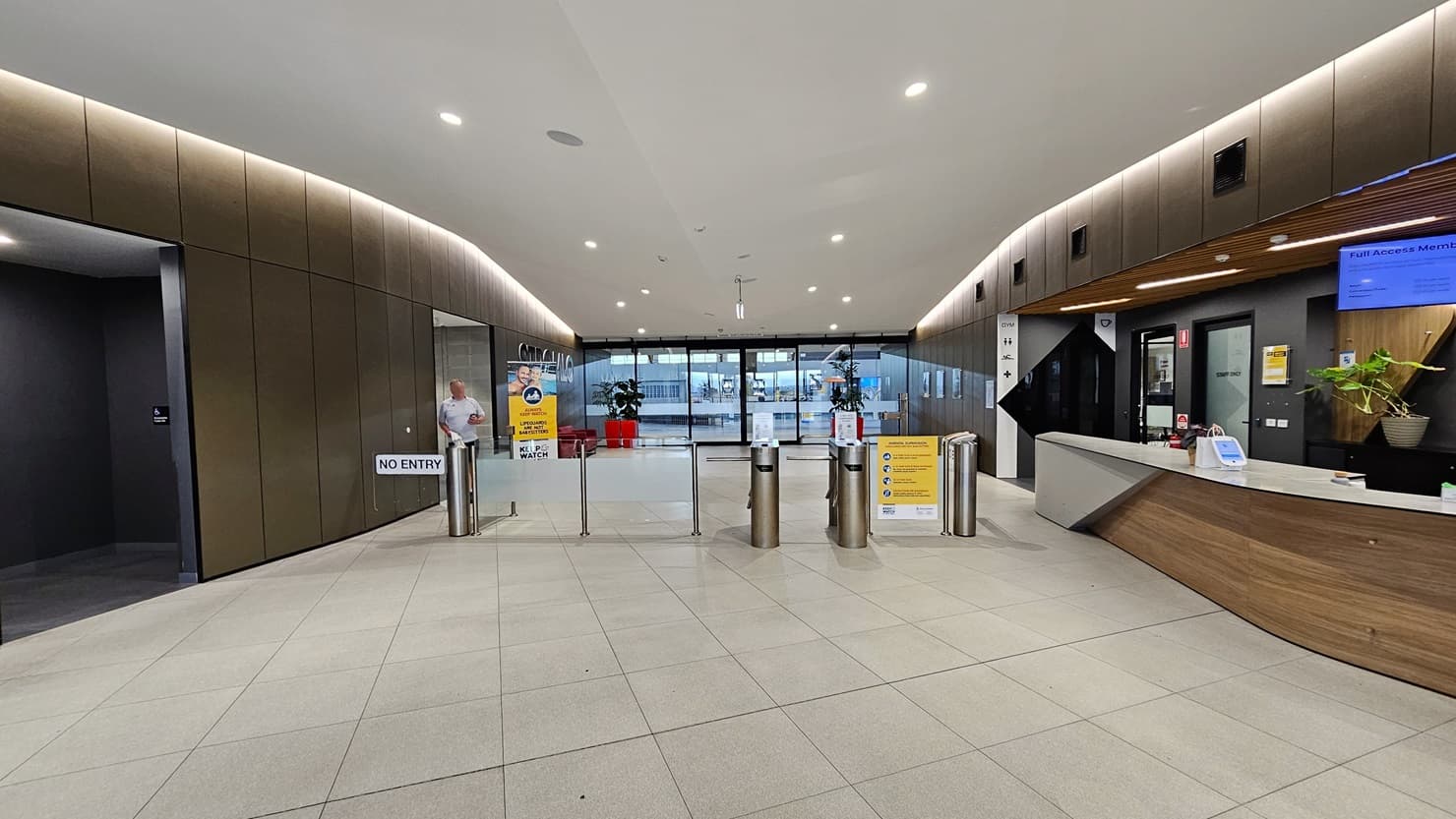 To the right is the reception desk, sporting a brown, white, and black front. A ceiling-mounted flat-screen TV is visible. In the middle is a turnstile gate. Further back, the main entrance is adorned with two red planter boxes and free-standing banners. On the left is an accessible bathroom. The reception desk extends into the corridor, leading to a seating area with chairs and tables on the right. The ceiling features a combination of white and brown with scattered LED lights.