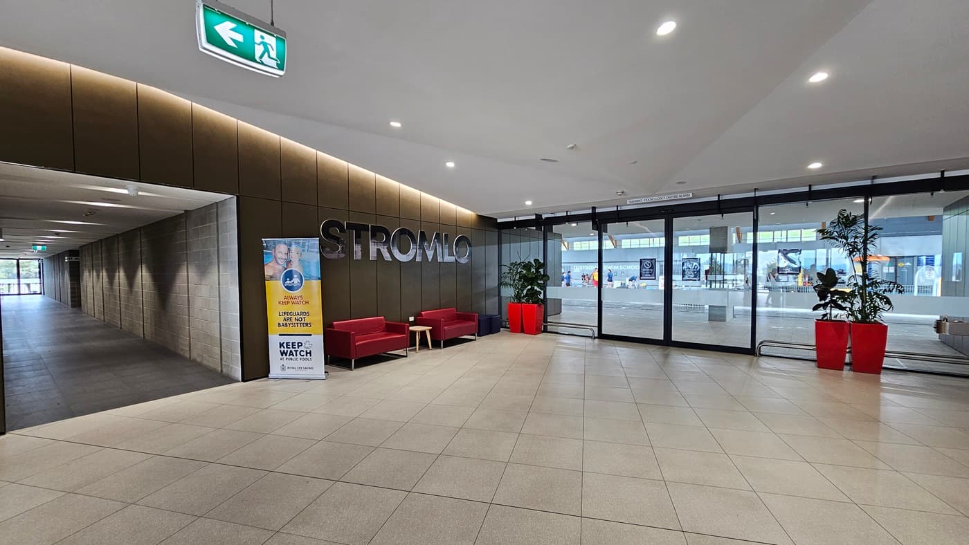 To the left is a corridor with grey brick walls. In the middle, red couches are arranged against the brown wall, with the facility's name on top. A retractable banner is positioned next to it. To the right, a glass door entry equipped with banners serves as the entry to the aquatic facility. Large red planter boxes with indoor plants are placed against the wall. The floor is white with LED lights, and grey tiles cover the floor.