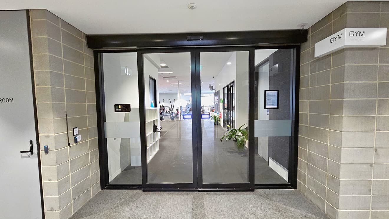 The entrance to the Gymnasium is through an automated glass sliding door adorned with posters and a sticker accent. A “Gym” sign is affixed to the upper left wall. The exterior wall is grey bricks, and the floor is tiled in grey. A grey door is visible on the left. The ceiling is uniformly white. Inside, a storage rack is located near the door.