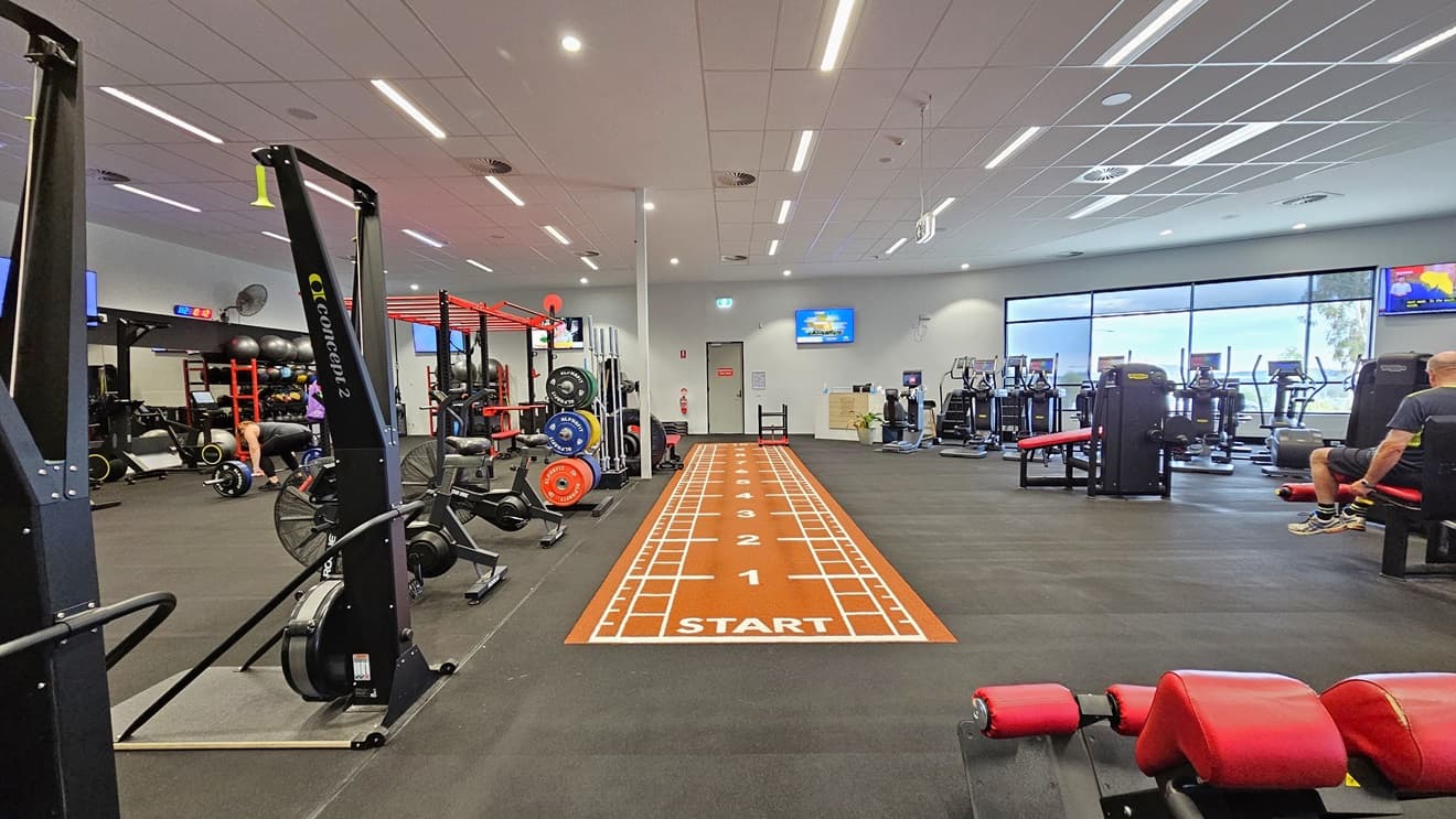 Inside the Gym, the left side houses Smith Machines, SkiErgs, Spin Machines, and free weights. In the middle, there is an orange sprint floor. Additional machines on the right include bench press, stair mill, and treadmills. Flatscreen TVs are mounted on the walls, and a glass wall surrounds the right side. The floor has black rubber tiles, and the ceiling features a white porous design with air conditioning and fluorescent lights.