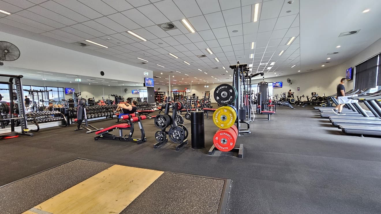 Inside the Gym, the free weights section and a Smith Machine are on the right. In the middle, there's a bench press, incline press, and Lat Pulldown machines. On the right, you'll find treadmills and stationary bikes, with additional equipment positioned behind. The floor is covered with black rubber tiles, with a brown accent and the ceiling features a white porous design with air conditioning and fluorescent lights.