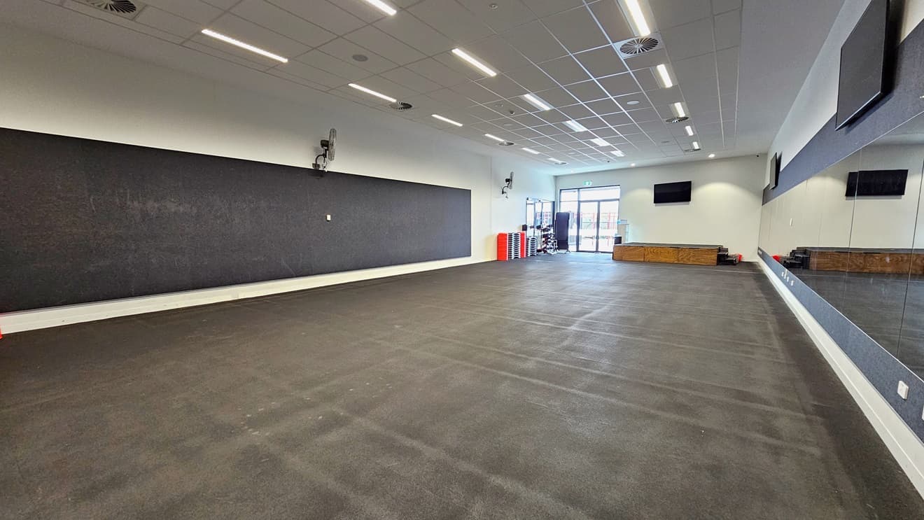 In the Fitness Room, a blackboard is on the left, and an expansive mirror is on the right. Flatscreen TVs are mounted on the upper middle and right walls, with wall fans installed on the upper left. Fitness equipment is stacked in the left corner near the glass door. The floor is covered with black rubber tiles, and the ceiling is white with fluorescent lights.