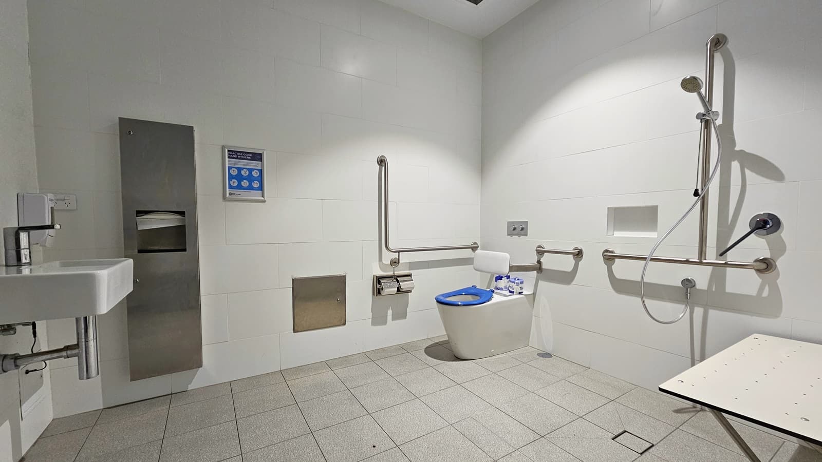 Inside the Accessible Bathroom, a toilet bowl with hand rails and a tissue dispenser is placed in the corner. To the right, a shower with handrails and a foldable bench is located. On the left, there's a washbasin next to a tissue and liquid soap dispenser. Grey tiles cover the floor, and the walls and ceiling are white. A poster is affixed to the right wall.