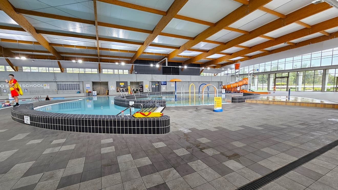 The entryway to the Indoor Aquatic Center features an automated glass sliding door adorned with three posters and a sticker accent. A sign is attached to the upper left wall. The exterior wall is constructed of grey bricks, while the floor is covered with grey tiles. The ceiling is uniformly white. Posters are displayed on the glass wall.