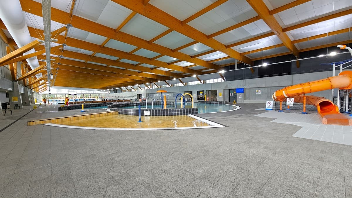 The children's splash park and leisure pool feature a beach entry, water sprays, geysers, and waterfalls. An orange slide is positioned on the right. Grey tiles surround the pool deck, and free-standing safety warnings and banners are placed on the slides. Plastic chairs are arranged on the left. The ceiling is white with wooden beams and large aircon vents. Additionally, a pool staff member is present.