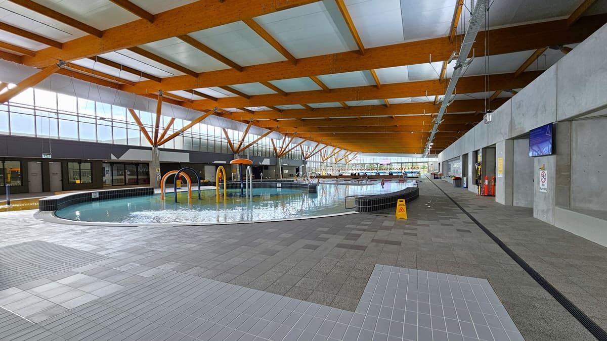 The children's splash park and leisure pool feature a beach entry, water sprays, geysers, and waterfalls. The pool has a raised deck with small black tiles, surrounded by grey checkered tiles. A yellow safety sign is visible, and a flat-screen TV is mounted on the right wall. The ceiling is embellished with wooden beams. A pool staff member is stationed beside the pool, and a glass wall surrounds the right section.