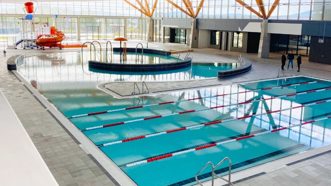 The training pool features handrails and floating ropes. Connected to it is the children's splash park and leisure pool, equipped with a beach entry, water sprays, geysers, and waterfalls. The pool has a raised deck with small black tiles, bordered by grey checkered tiles. Furthermore, a slide with a tipping bucket is positioned near the glass wall. People are standing beside the pool on the right.