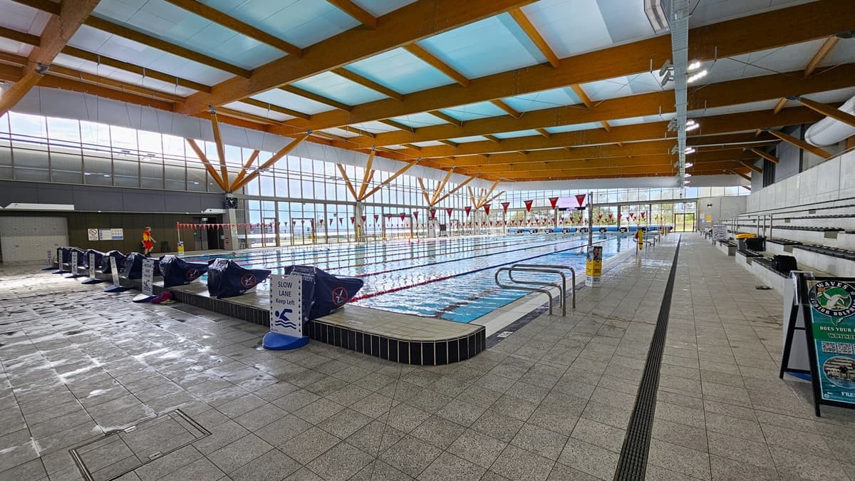 The 50m Heated Swimming Pool has handrails, floating ropes, and banners. Diving boards, wrapped with blue covers, are aligned on the raised pool deck on the left. Free-standing safety banners are placed on the pool deck. On the right, there is tiered seating with additional free-standing banners. The ceiling is adorned with wooden beams and floodlights. The floor is covered with grey tiles, featuring an overflow grid. A glass wall surrounds the pool.