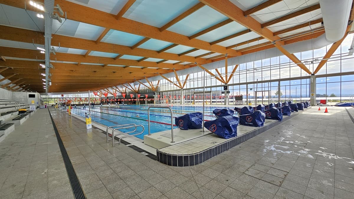 The 50m Heated Swimming Pool features handrails, floating ropes, and orange banners. Diving boards, covered in blue, are aligned on the raised pool deck on the right. Safety cones and banners are visible on the pool deck. The ceiling is adorned with wooden beams and floodlights. The floor is covered with grey tiles, including an overflow grid. The pool is surrounded by a glass wall.
