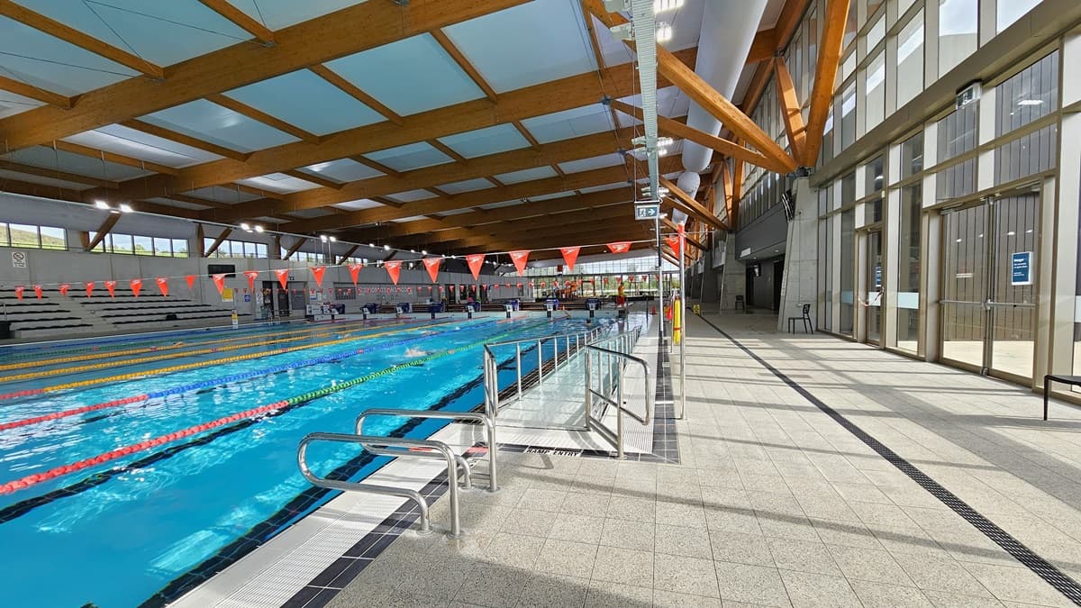 The 50m Heated Swimming Pool includes a ramp with handrails, floating ropes, and orange banners. At the pool's end, diving boards, covered in blue, align with the raised pool deck. To the right, a glass wall and door are present. The ceiling features wooden beams, large aircon vents, and floodlights. The floor is tiled in grey, incorporating an overflow grid. To the right is a glass wall and a glass door.