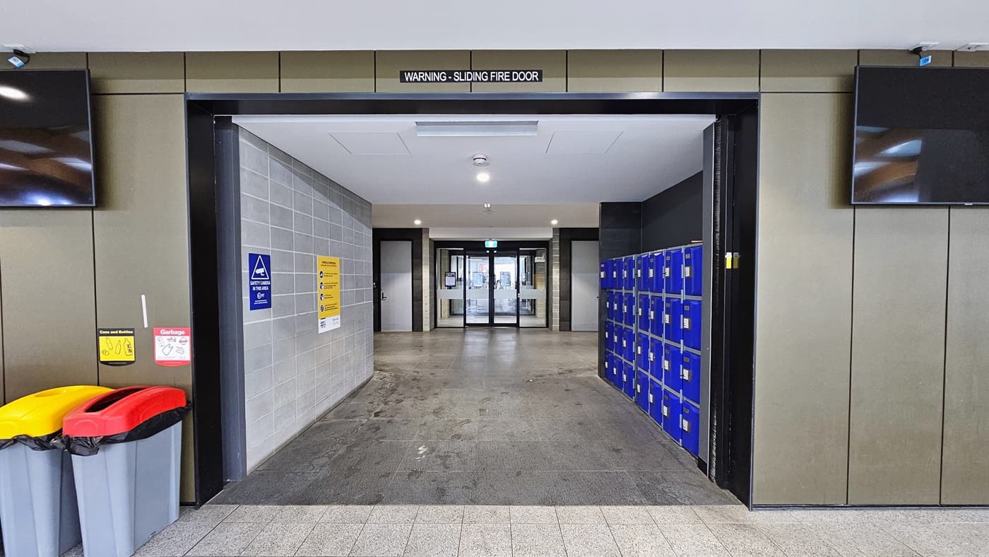 Blue lockers are situated on the right, while the grey brick wall opposite displays posters and safety signs. Outside, two rubbish bins with segregation signs are placed on the left, beneath a flatscreen TV. Another flatscreen TV is mounted on the right wall. The floor transitions from light to dark grey, and the ceiling is uniformly white. Further in, there is a glass door entry.
