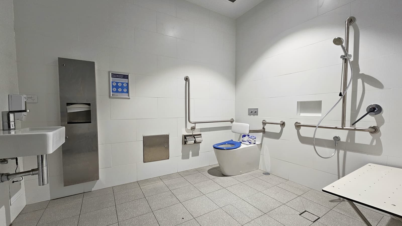 Within the accessible bathroom, a toilet bowl equipped with hand grabs and a tissue dispenser is positioned in the corner. To its right, there is a shower with handrails, accompanied by a foldable bench. On the left is a washbasin, adjacent to a tissue and liquid soap dispenser. The floor is covered with grey tiles, and the walls and ceiling are white.