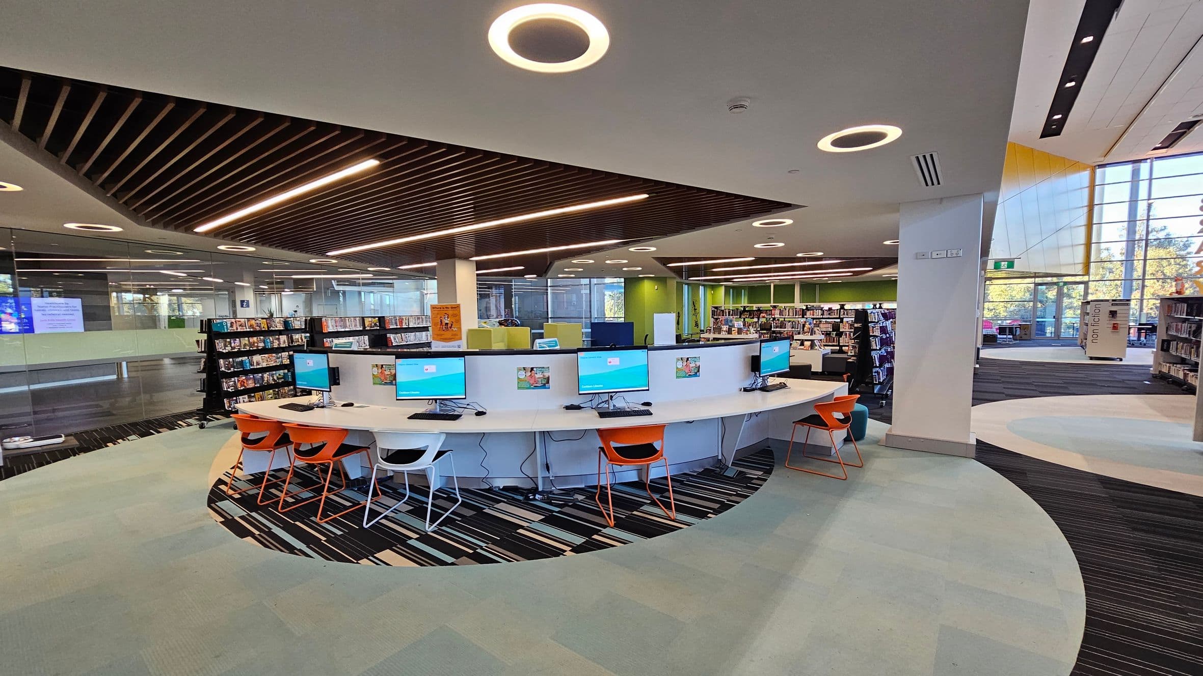 A modern library interior with a circular computer station in the centre, featuring several monitors and orange and white chairs. The floor has a mix of striped and solid-coloured carpet tiles. Bookshelves filled with books are visible in the background, along with a glass wall on the left. The ceiling has circular and linear lights, with a section of wooden slats. Large windows on the right allow natural light to enter, and a sign marked "non-fiction" is visible.