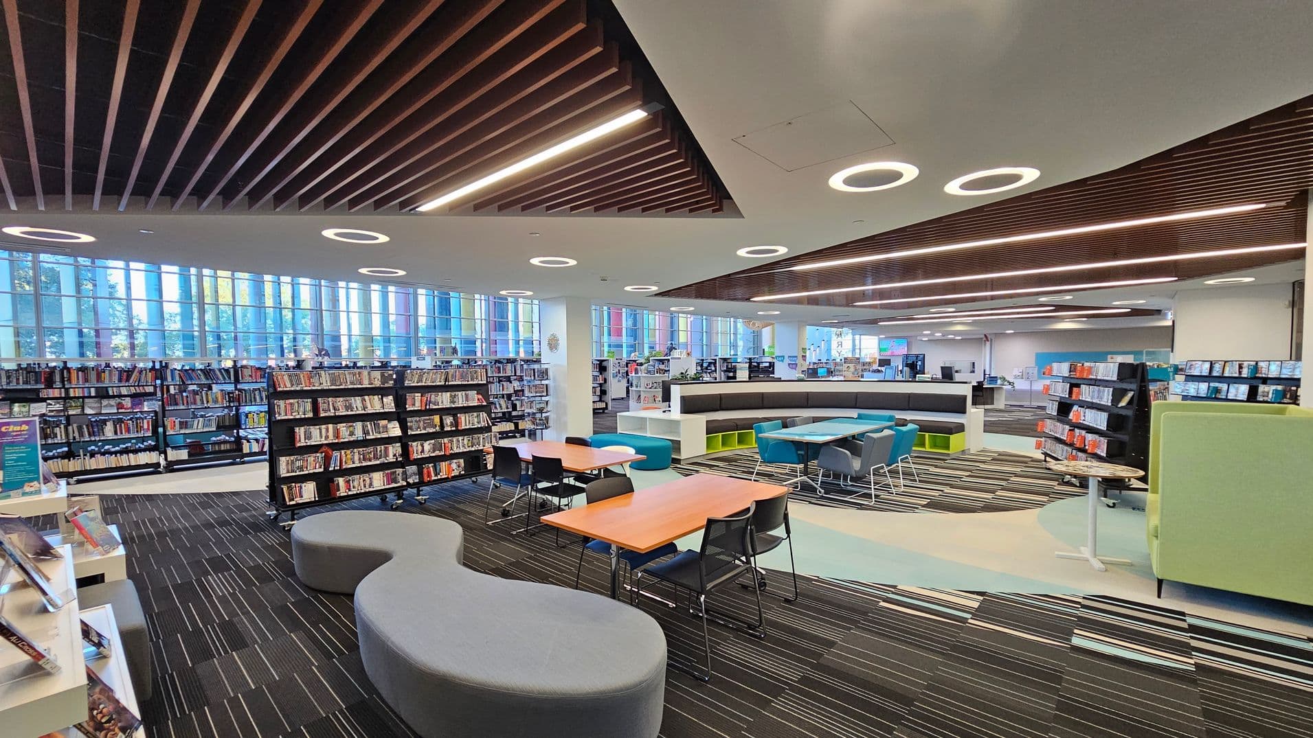 A modern library interior with a striped carpeted floor. On the left, there are bookshelves filled with books and DVDs. In the centre, there are wooden tables with black chairs and a grey, curved bench. On the right, more bookshelves and a light green high-backed chair are visible. The ceiling features wooden slats and circular lights. Large windows in the background allow natural light to flood the space.