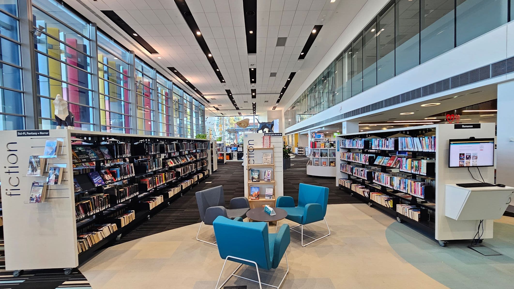 A modern library interior with a high ceiling and large windows on the left, featuring colourful vertical panels. Bookshelves line both sides, with a focus on fiction and romance sections. In the centre, there are four chairs in blue and grey around a small round table. The floor is carpeted in a mix of dark and light patterns. A computer station is visible on the right, and the upper level has glass panels. The space is well-lit with ceiling lights.