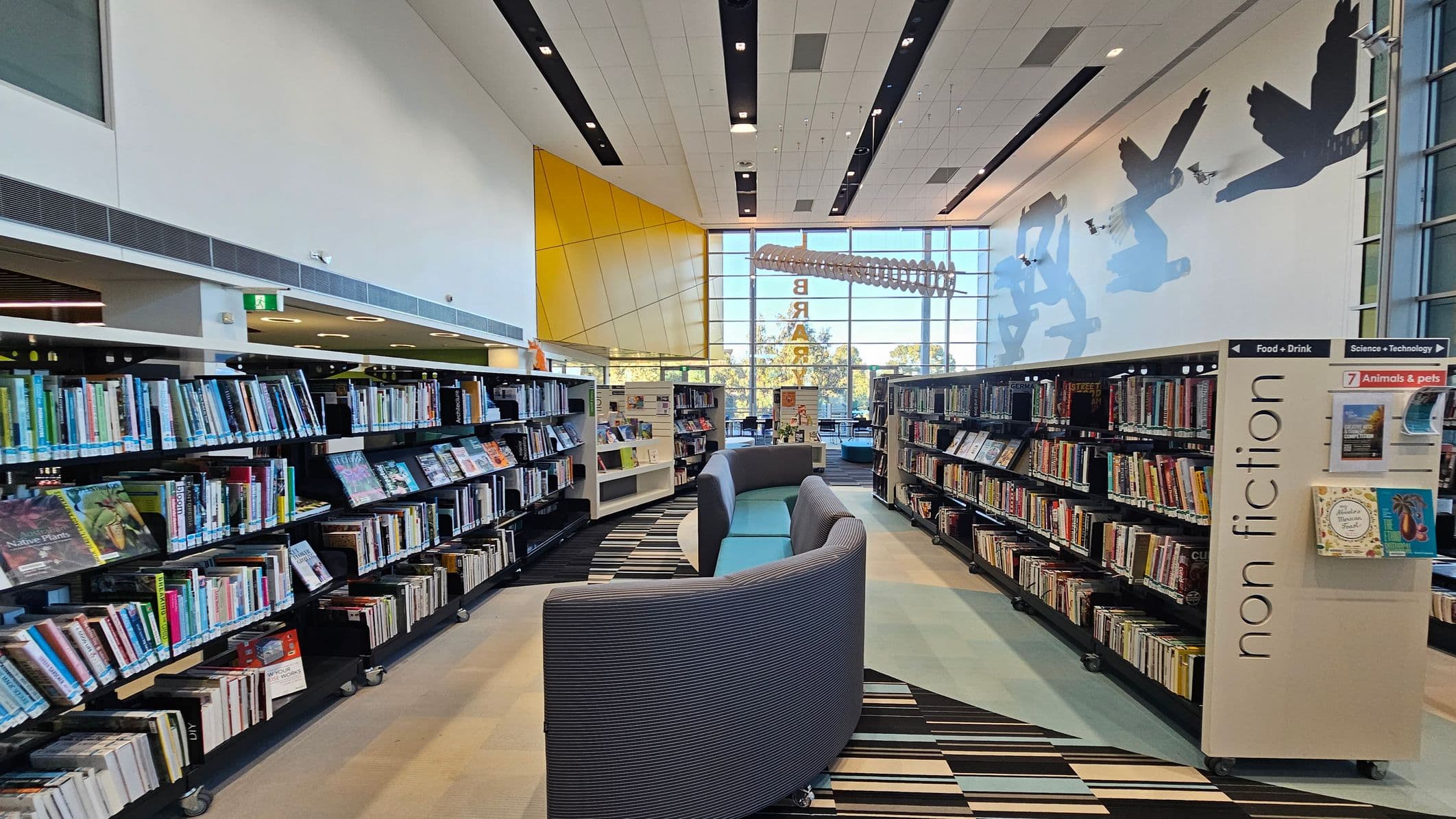 Bookshelves filled with books line both sides of a library aisle. On the left, the shelves are labelled with various categories. On the right, a sign reads "non fiction" with sections for "Food + Drink" and "Science + Technology". In the centre, curved grey seating is placed on a striped carpet. The background features large windows with a view of trees and a yellow wall. The ceiling is white with black panels and recessed lighting. Silhouettes of birds are painted on the right wall.