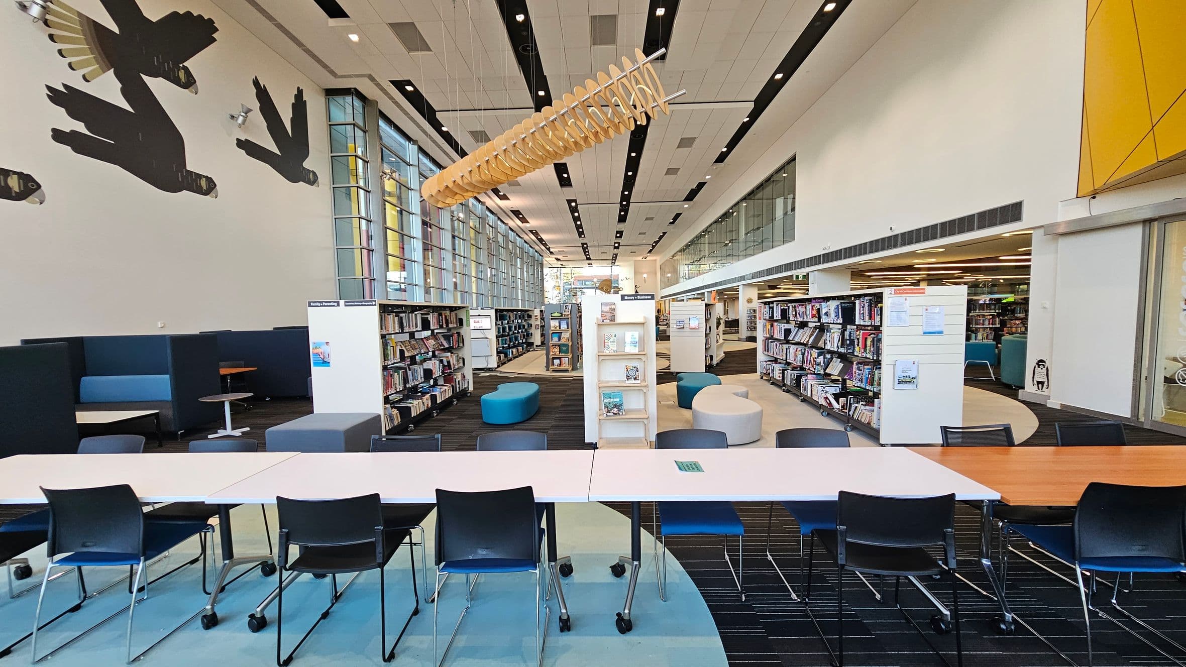 A modern library interior with a long table and chairs in the foreground. The left wall features large bird silhouettes and blue seating booths. The centre has bookshelves and a few blue and grey ottomans. The right side has more bookshelves and a yellow wall section. The ceiling is white with black panels and a unique wooden light fixture. Large windows line the left side, allowing natural light to fill the space.