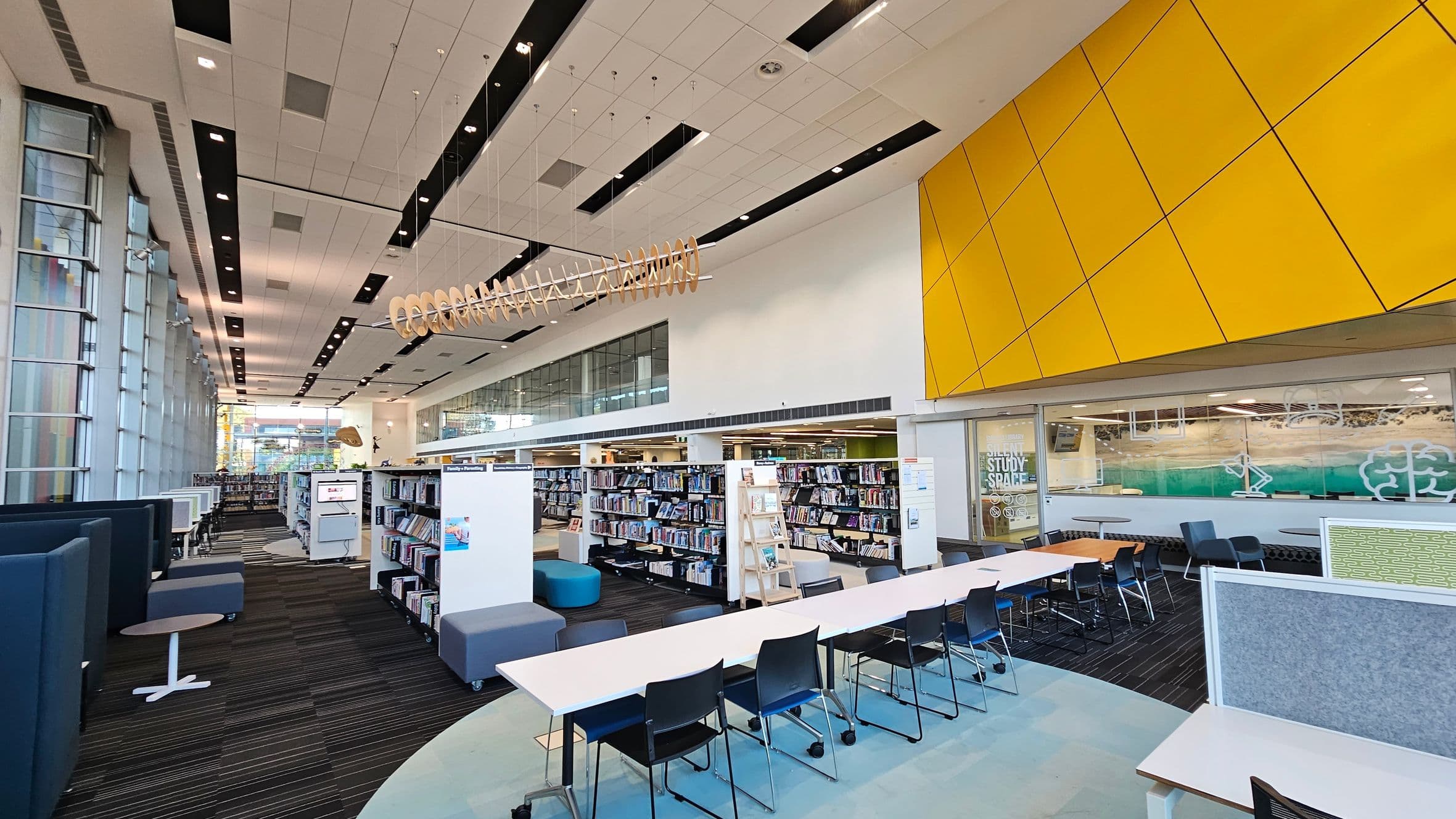 A modern library interior with a high ceiling featuring black and white panels. On the left, there are tall glass windows and a row of blue seating booths with small tables. In the centre, bookshelves are filled with books, and a long white table with black chairs is visible. On the right, a large yellow panelled wall is above a glass-walled study space. The floor is carpeted with a striped pattern, and a unique wooden sculpture hangs from the ceiling.