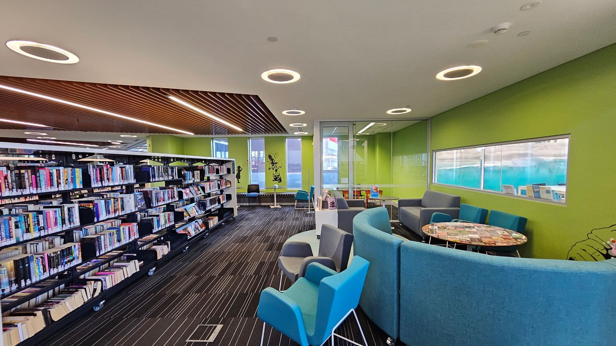 A modern library interior with a green wall on the right featuring a long horizontal window. In the foreground, there are blue and grey armchairs around a circular table. On the left, there are bookshelves filled with books. The ceiling has circular light fixtures and a section with wooden slats. The floor is carpeted with a striped pattern. In the background, there are more seating areas and a glass-walled room.