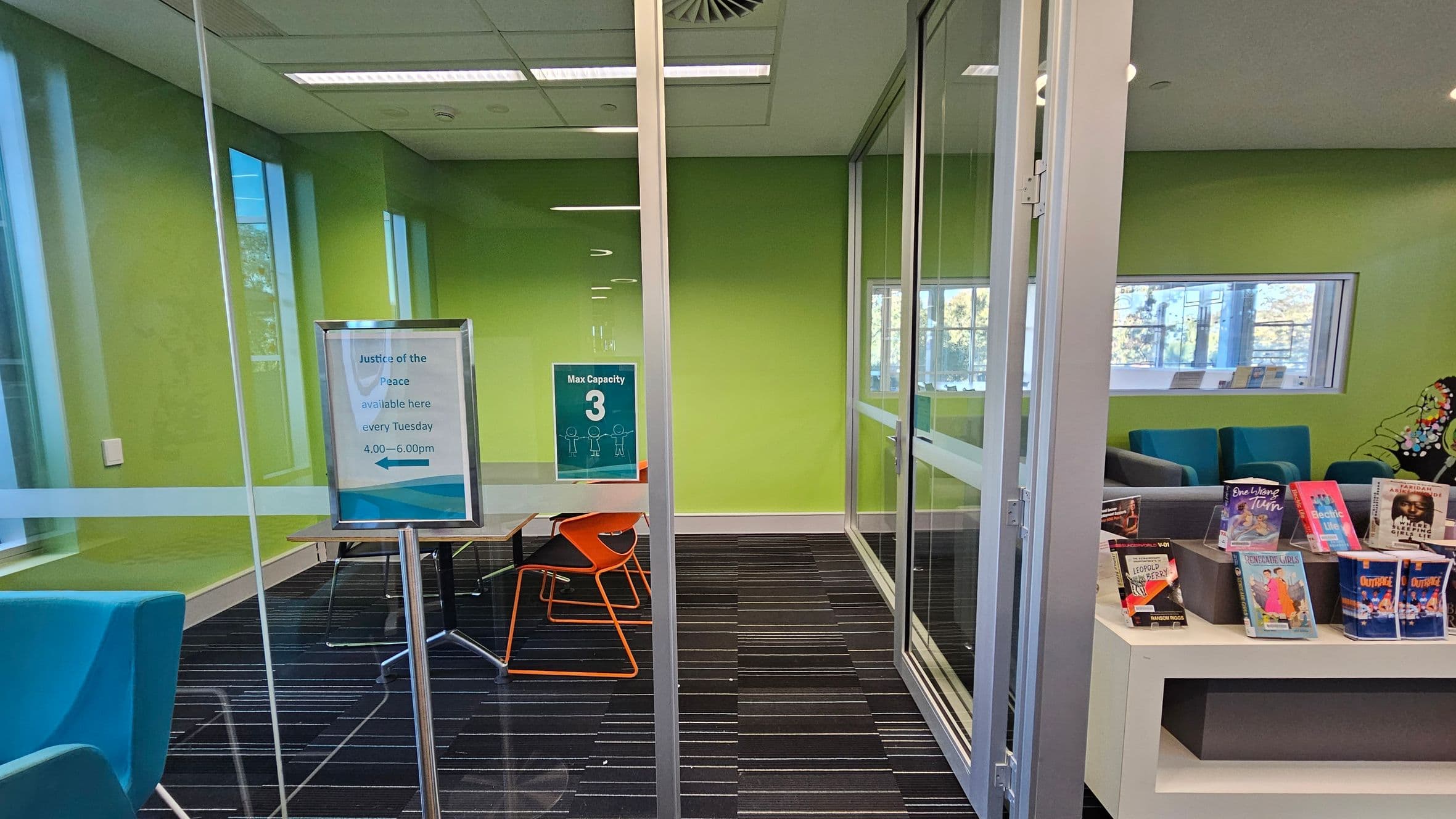 A room with green walls and a glass partition. On the left, a sign on a stand reads "Justice of the Peace available here every Tuesday 4.00—6.00pm." Below it, a table with orange chairs. On the right, a sign indicates "Max Capacity 3." In the foreground, a shelf displays various books. The floor is dark with striped patterns, and the ceiling has recessed lighting and a vent. A window in the background shows trees outside.