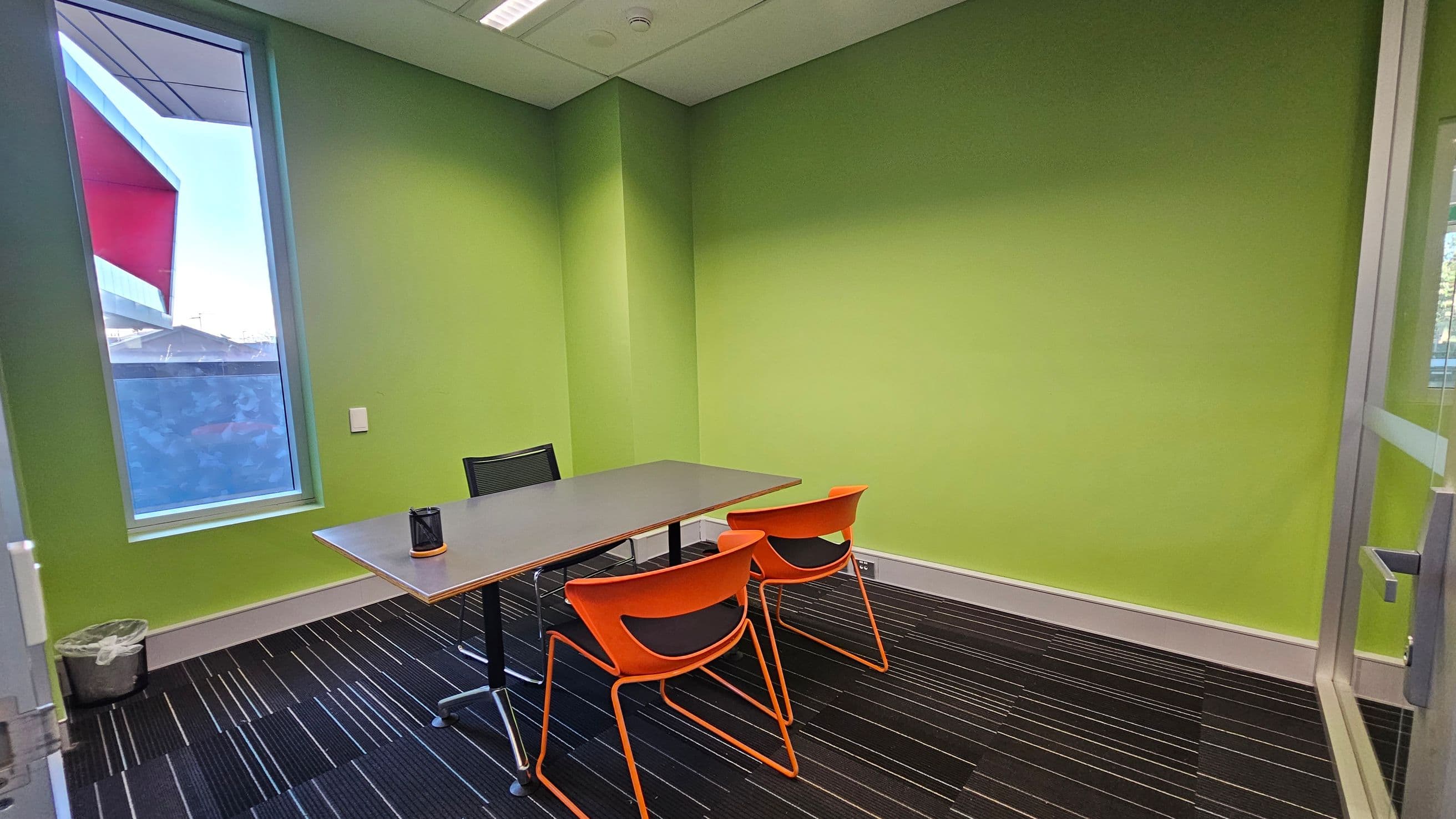 A small meeting room with bright green walls. On the left, a tall window with a view of a building and a red awning. Below the window, a small waste bin. In the centre, a rectangular table with a black chair on one side and two orange chairs on the other. The floor is covered with a dark carpet featuring thin, colourful stripes. The ceiling has recessed lighting.