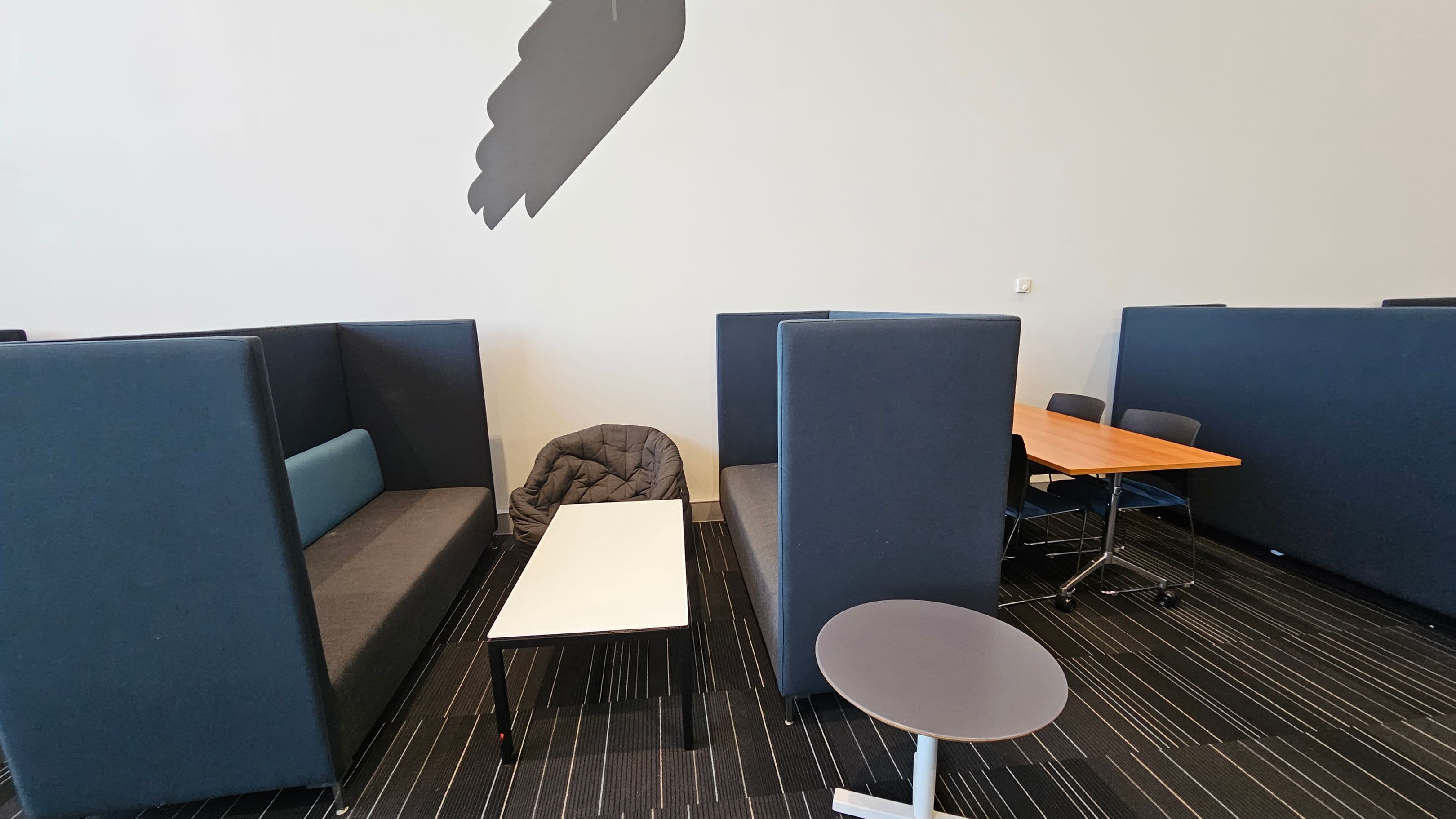 On the left, there are two high-backed blue and grey sofas facing each other with a white rectangular table in between. A grey cushioned chair is placed next to the table. On the right, there is a wooden table with chairs around it, partially visible. The floor is covered with a dark carpet featuring thin, colourful stripes. The wall is plain white with a shadow of an abstract shape cast on it. A small round table is in the foreground.