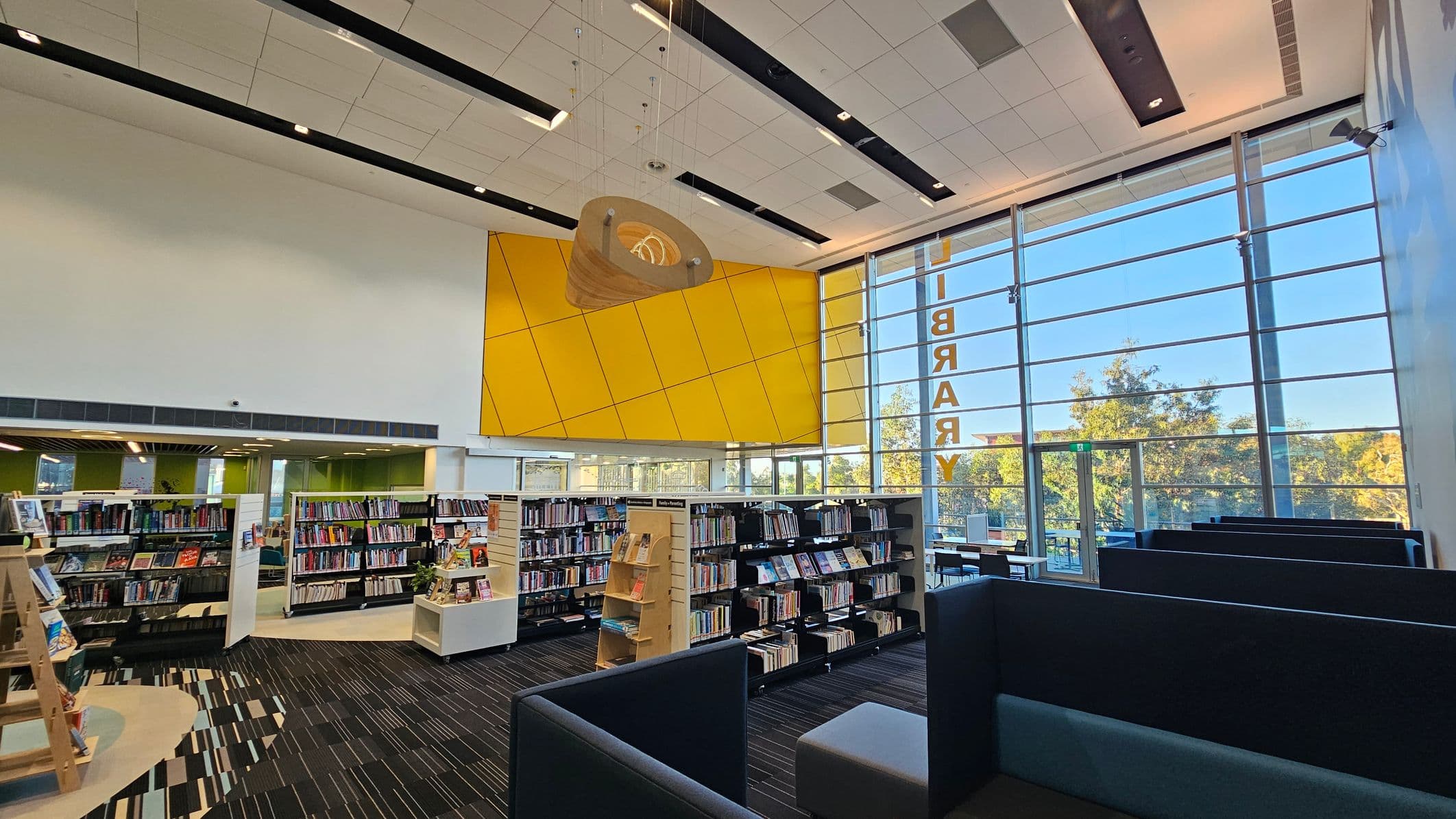 Modern library interior with a high ceiling and large windows on the right, allowing natural light to illuminate the space. Bookshelves filled with books are in the centre and left, with a mix of wooden and metal shelving. A striking yellow geometric wall feature is visible above the shelves. On the right, there are dark-coloured seating booths. The floor is carpeted with a striped pattern, and the ceiling has a grid design with recessed lighting. Trees are visible through the windows.