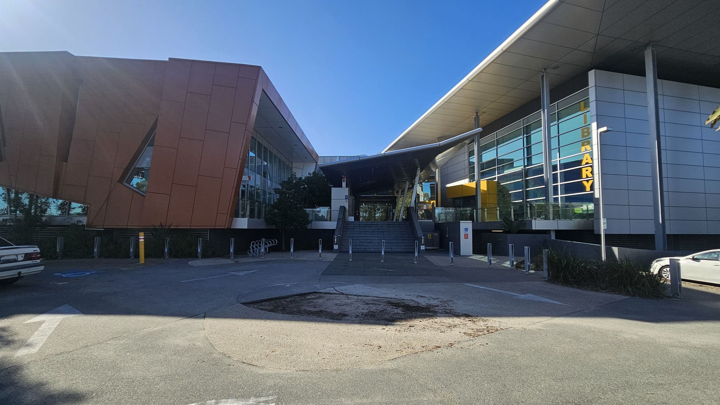 A modern building with a unique architectural design. On the left, there is a structure with angular, reddish-brown panels and a large, slanted window. In the centre, a set of stairs leads up to the entrance, flanked by glass railings and greenery. On the right, a building with a sleek, silver facade and large windows displays the word "LIBRARY" in vertical yellow letters. The foreground features a paved area with a circular patch of exposed ground.
