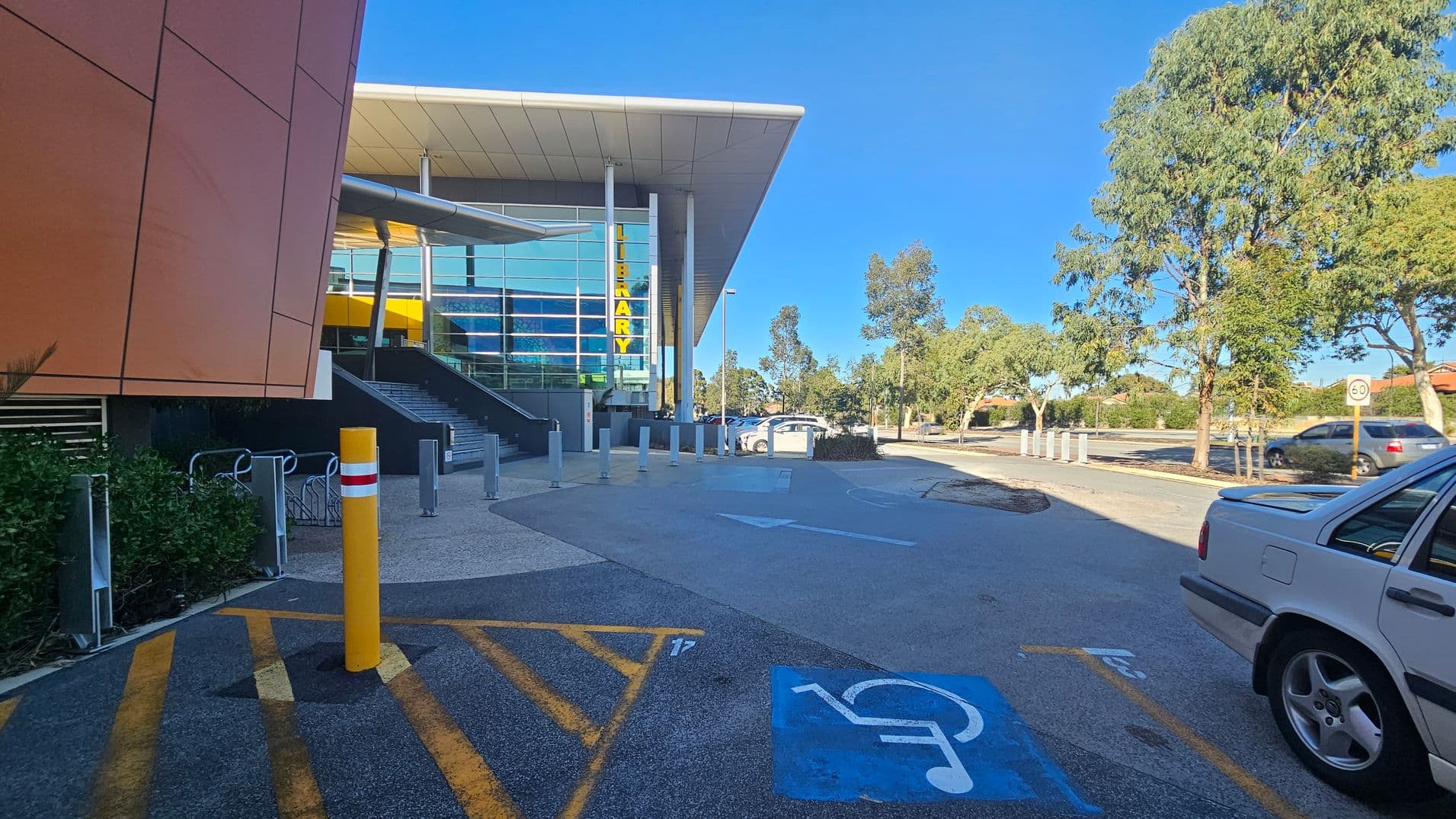 A modern building with a large glass facade and a sign reading "LIBRARY" in yellow letters is on the left. The entrance features stairs and a ramp. In the foreground, there is a marked disabled parking space with a blue symbol. A yellow bollard and bike racks are nearby. On the right, a silver car is parked, and several trees line the road. The sky is clear, and a speed limit sign is visible in the background.