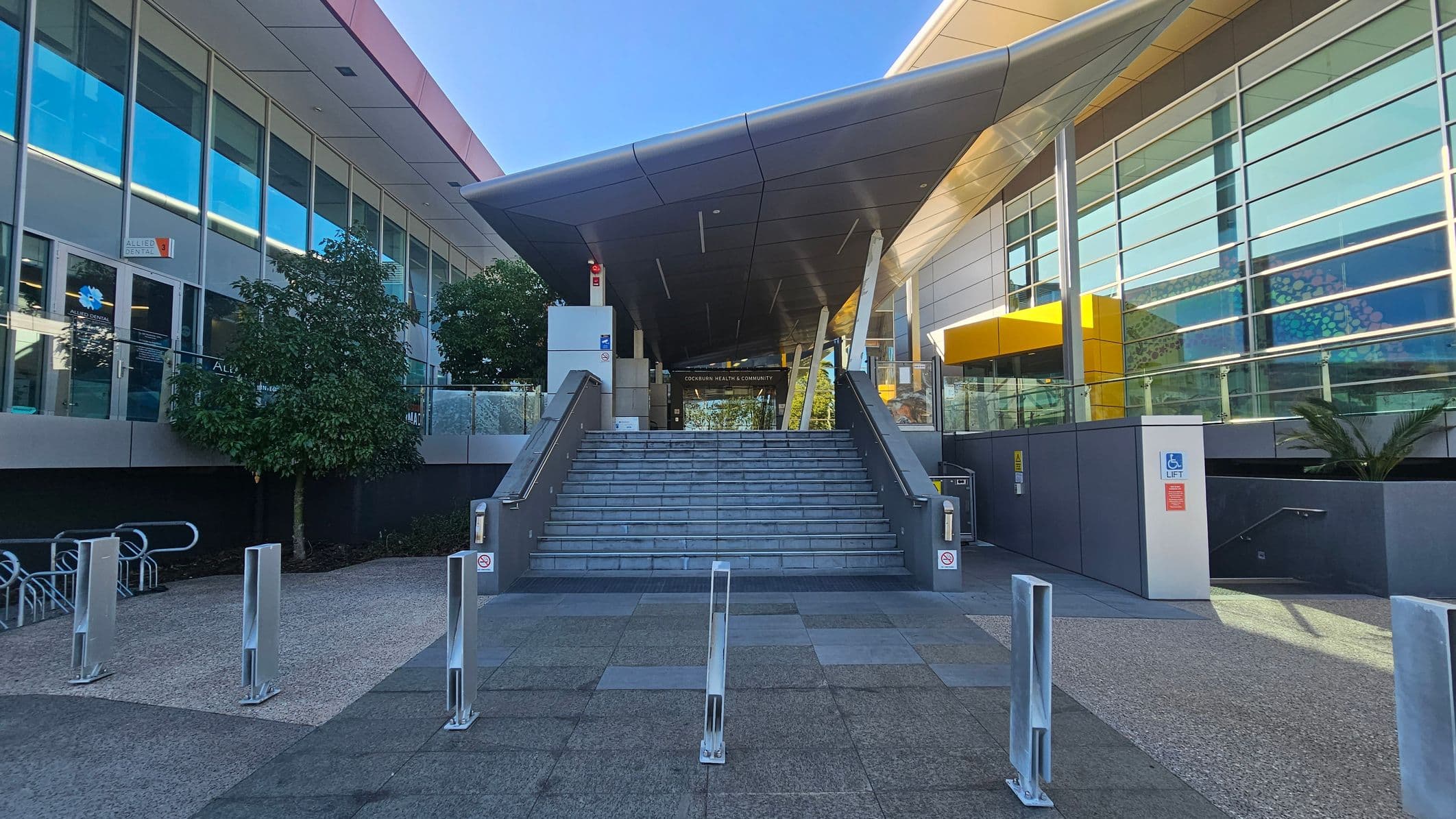 A modern building entrance with a wide staircase leading up to a covered area. On the left, there are large glass windows and a sign for "Allied Dental" near some trees. On the right, a series of horizontal windows with a yellow accent above a doorway. The foreground features a paved area with metal bollards. The ceiling above the entrance is angular and supported by slanted columns.