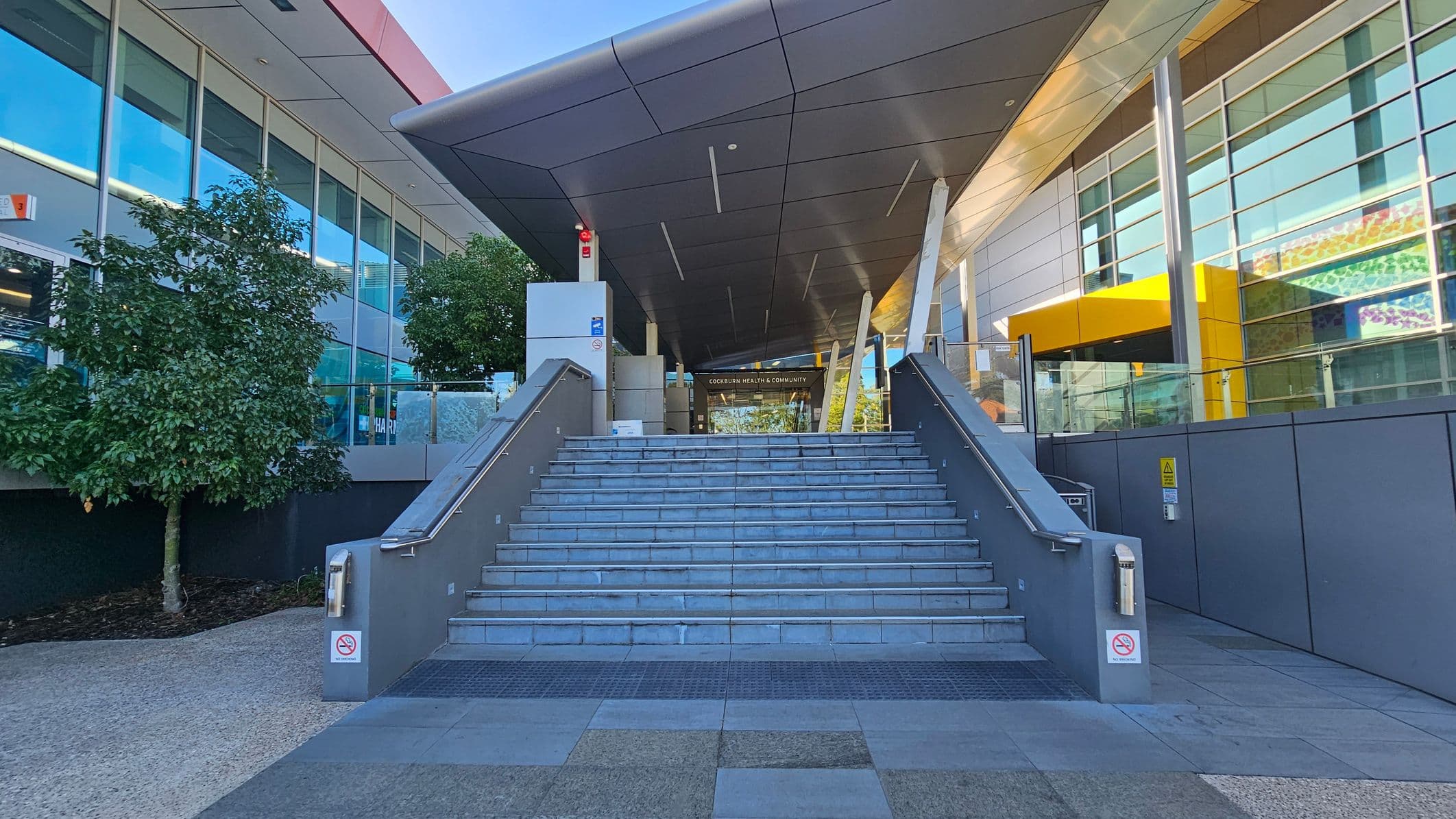 Wide stone steps lead up to a modern building entrance. On the left, there are large windows and a tree. On the right, a yellow architectural feature and more windows. The ceiling is angular and metallic, with supporting beams. The floor is paved with grey tiles. Signs and handrails are present on the stairs.