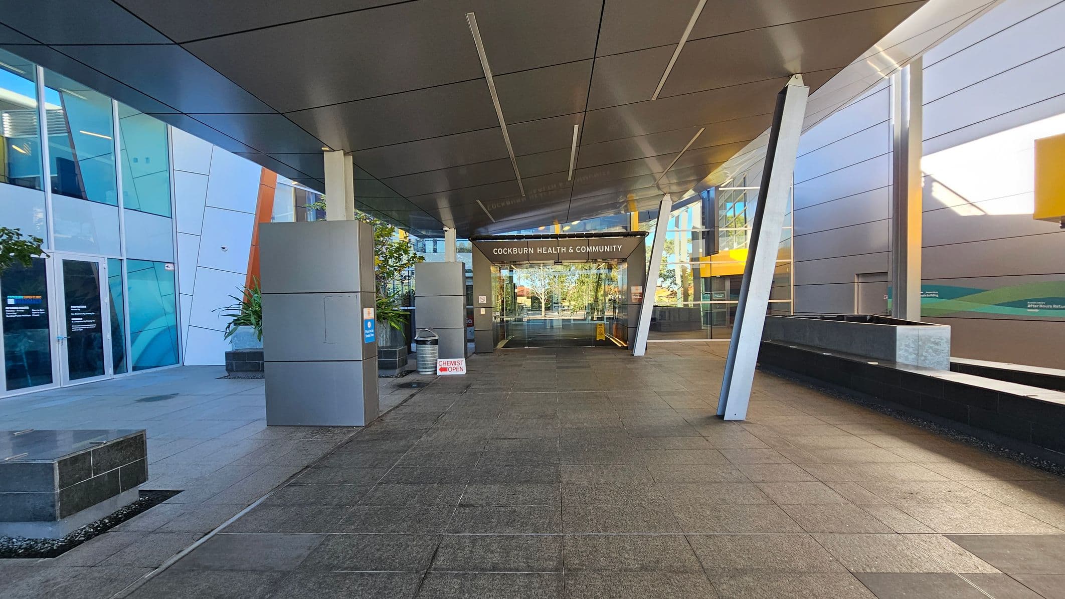 A modern building entrance with a grey tiled floor and a sleek, angular ceiling. On the left, there are large glass windows and a door, with some potted plants nearby. A sign indicates a chemist is open. In the centre, the entrance to "Cockburn Health & Community" is visible, with glass doors reflecting the outside. On the right, there is a long, low planter box and more glass panels. The structure features metal beams and a contemporary design.