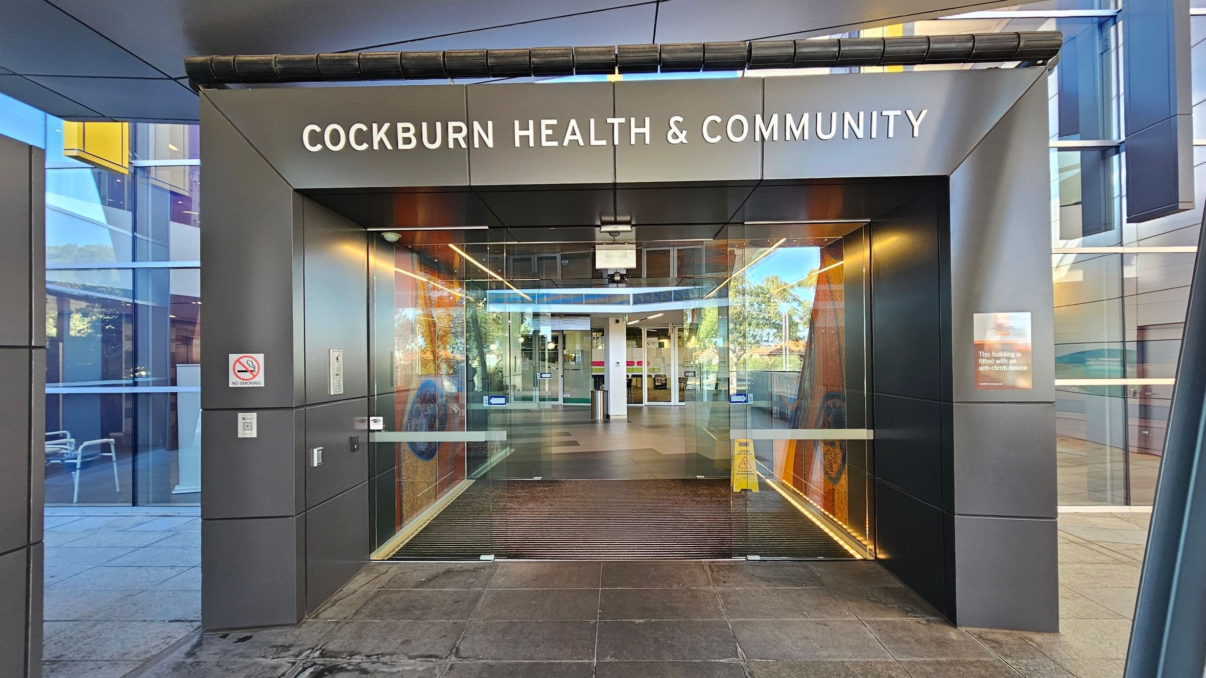 Entrance to a modern building with a sign reading "Cockburn Health & Community" above. The entrance features large glass doors and a dark grey frame. A "No Smoking" sign is visible on the left side, and a notice about an anti-climb device is on the right. Inside, a wet floor sign is placed on the tiled floor. The exterior walls are made of reflective glass panels, and there are some chairs visible through the glass on the left.