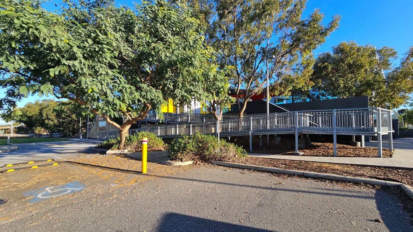 A paved area with a disabled parking space marked in blue on the left. A yellow bollard stands nearby. A tree with dense green foliage is on the left, casting shadows on the ground. In the centre, a metal railing encloses a raised walkway leading to a building with colourful panels in yellow, white, and red. More trees with sparse leaves are in the background, and the ground is covered with wood chips and small shrubs.