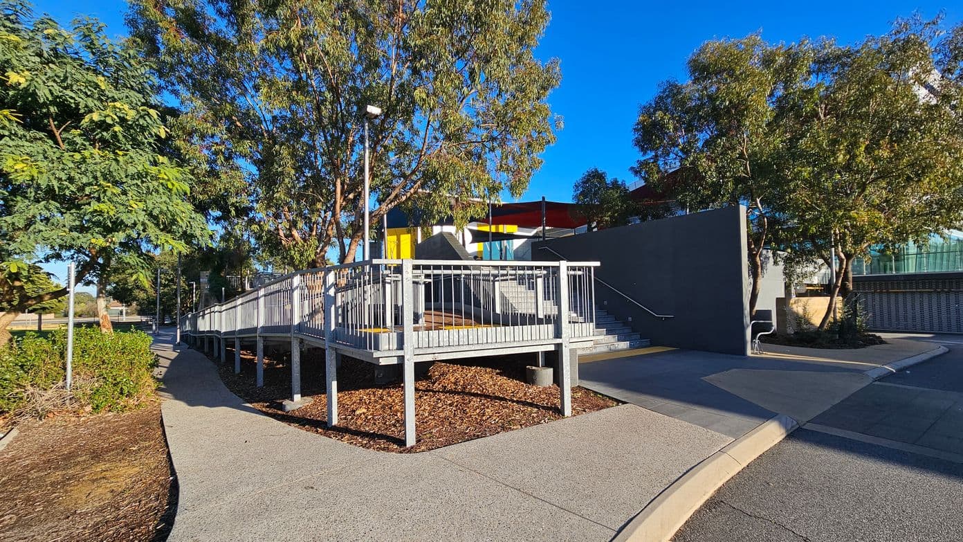 A modern outdoor area with a ramp and stairs leading to a building. The ramp has metal railings and is surrounded by trees and shrubs. The building features a yellow door and large windows. The ground is covered with mulch and concrete paths. A dark wall is adjacent to the stairs, and a bench is visible near the trees. The sky is clear and blue.