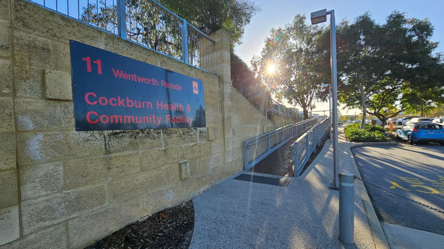 A stone wall on the left features a sign for "Cockburn Health & Community Facility" at "11 Wentworth Parade." A ramp with blue railings runs alongside the wall. On the right, a paved road with parked cars is visible, with a blue car in the foreground. Trees line the road, and sunlight filters through the branches, casting shadows on the ground. A tall streetlight stands near the ramp.
