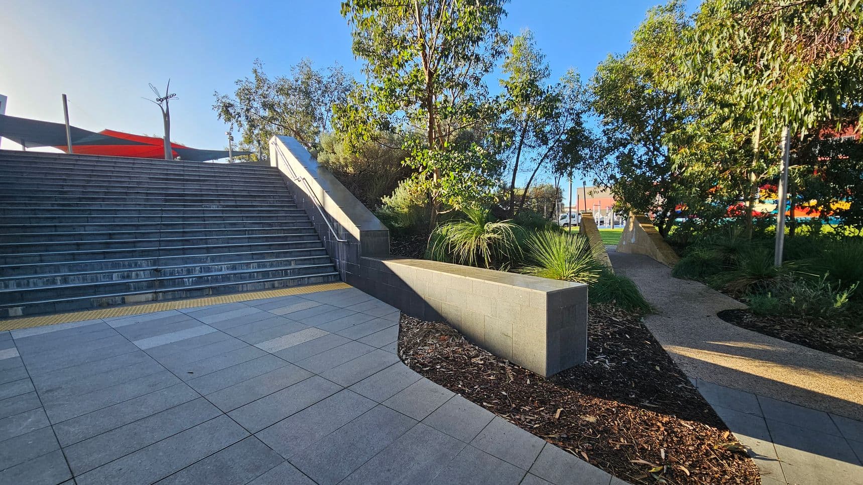 Wide stone steps on the left lead upwards, bordered by a low wall. A path on the right curves through a landscaped area with trees and shrubs. The ground is paved with large tiles, and there is a textured yellow strip at the base of the steps. In the background, colourful structures and a clear blue sky are visible.