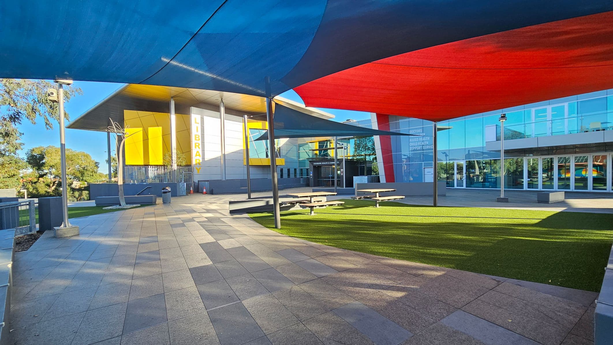 A modern building with a large sign reading "LIBRARY" on the left, featuring yellow and white panels. The foreground has a paved area with a patch of artificial grass and several picnic tables. Overhead, colourful shade sails in blue and red provide cover. The right side of the building has glass windows and doors, with signage indicating health services. Trees are visible in the background, and there are metal poles supporting the shade sails.