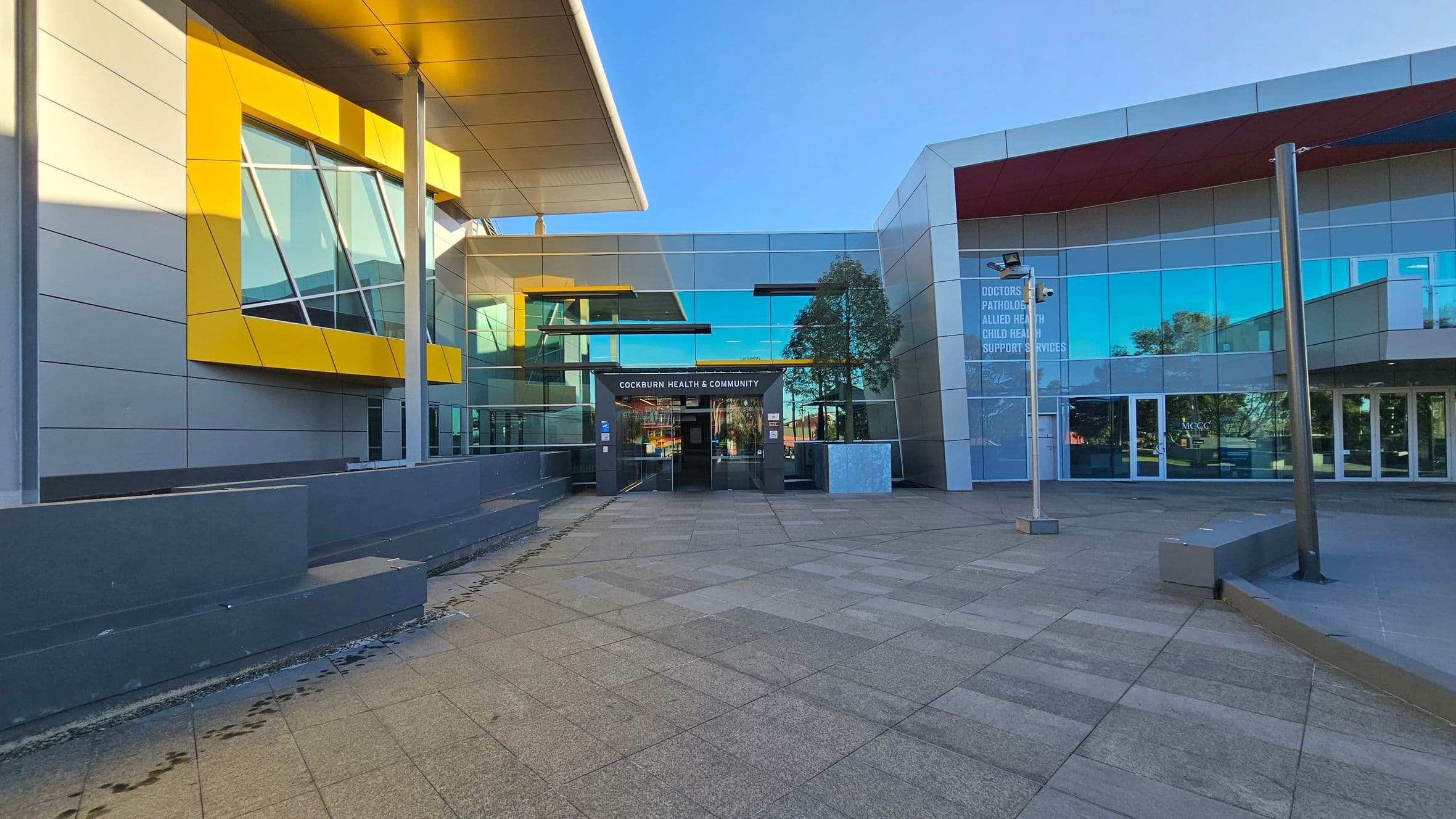 A modern building with a glass facade and angular design elements. On the left, there is a large yellow-framed window. The entrance in the centre has a sign above it. The right side features more glass panels with text indicating various services. The ground is paved with large tiles, and there are metal poles and a small tree near the entrance. The ceiling has a metallic finish with a slight overhang.