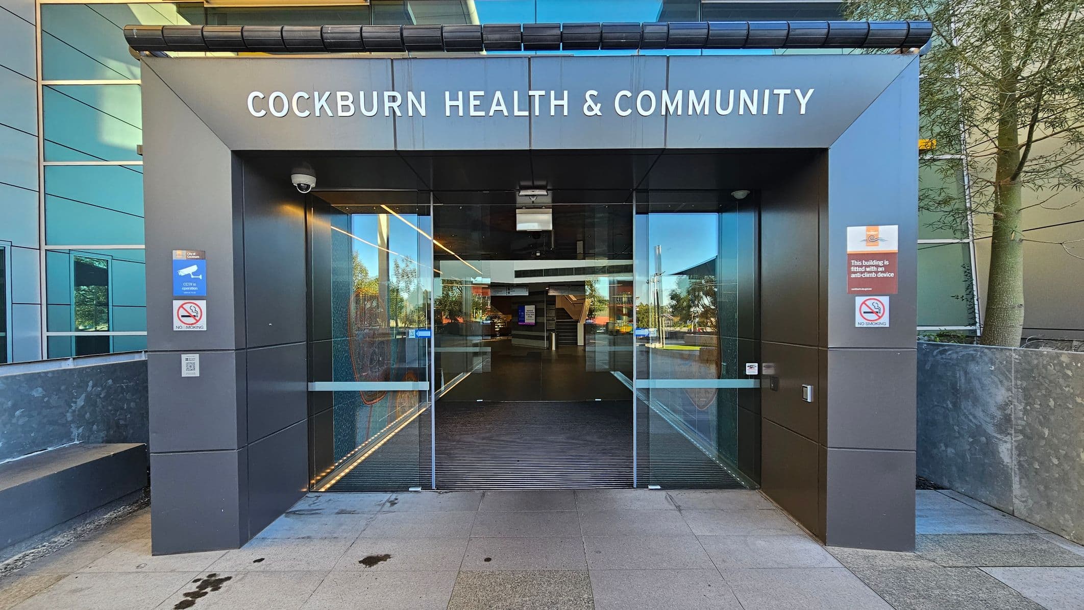Entrance to a modern building with "Cockburn Health & Community" written above. The entrance features large glass doors with reflections of trees and the surroundings. Signs on either side indicate CCTV surveillance and no smoking. The exterior walls are clad in dark panels, and there is a tree to the right. The floor is paved with light-coloured tiles, and the ceiling inside is visible through the glass, showing a sleek, contemporary design.