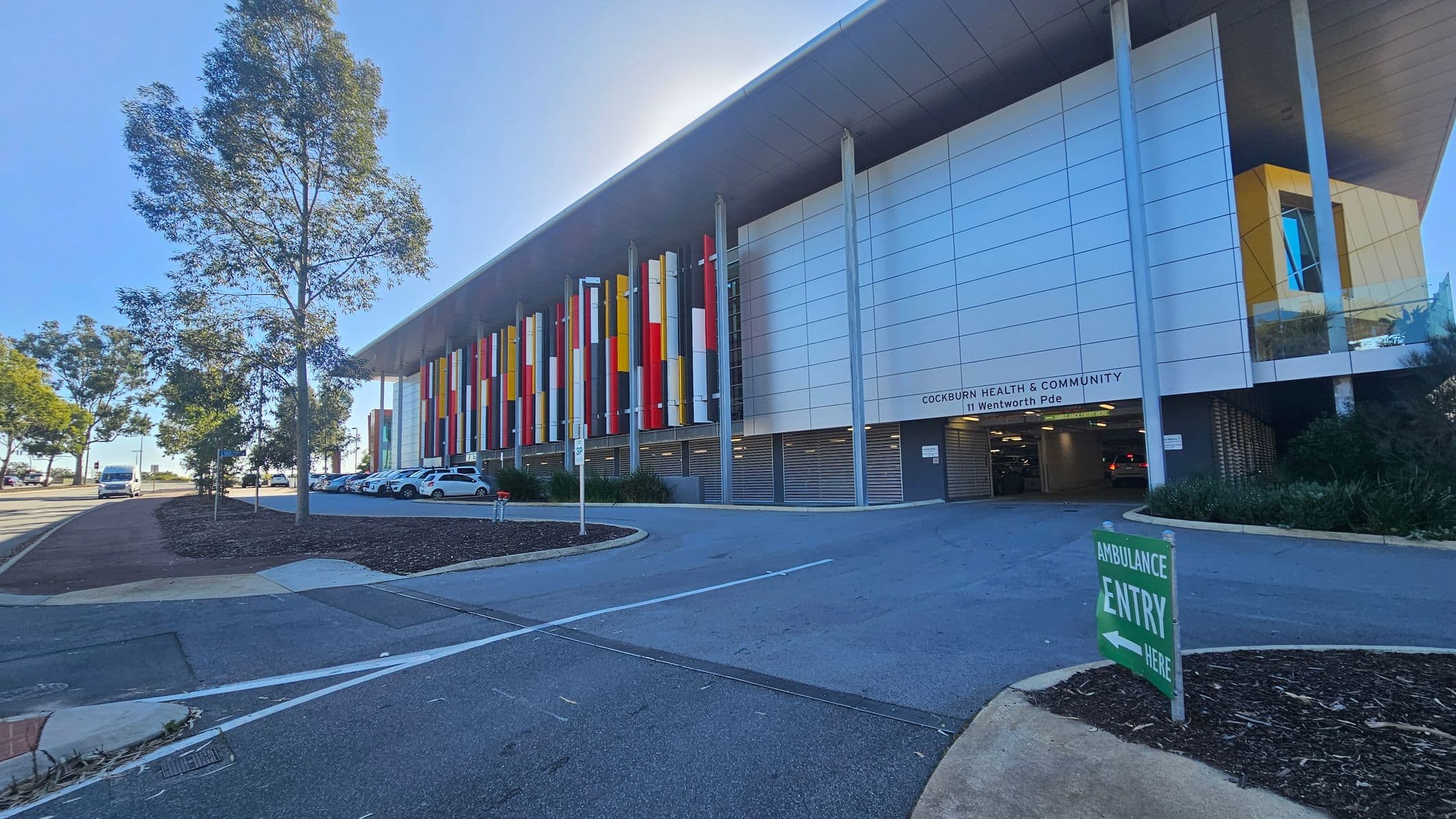 A modern building with a large overhanging roof and a facade featuring vertical panels in red, yellow, and white. The entrance is marked "Cockburn Health & Community" with a driveway leading to a parking area. Several cars are parked in front. A green sign on the right indicates "Ambulance Entry." Trees line the left side of the image along a pathway, and the ground is covered with a mix of pavement and landscaped areas.